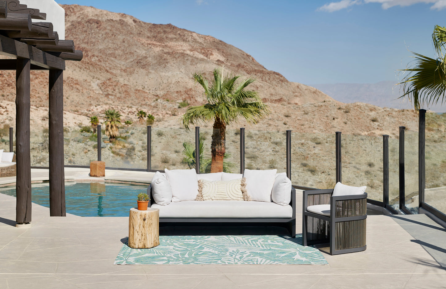 Outdoor scene with a cream sofa, dark chairs, a wooden stump table, and a teal palm-print runner rug beside a pool & desert hills in the background.