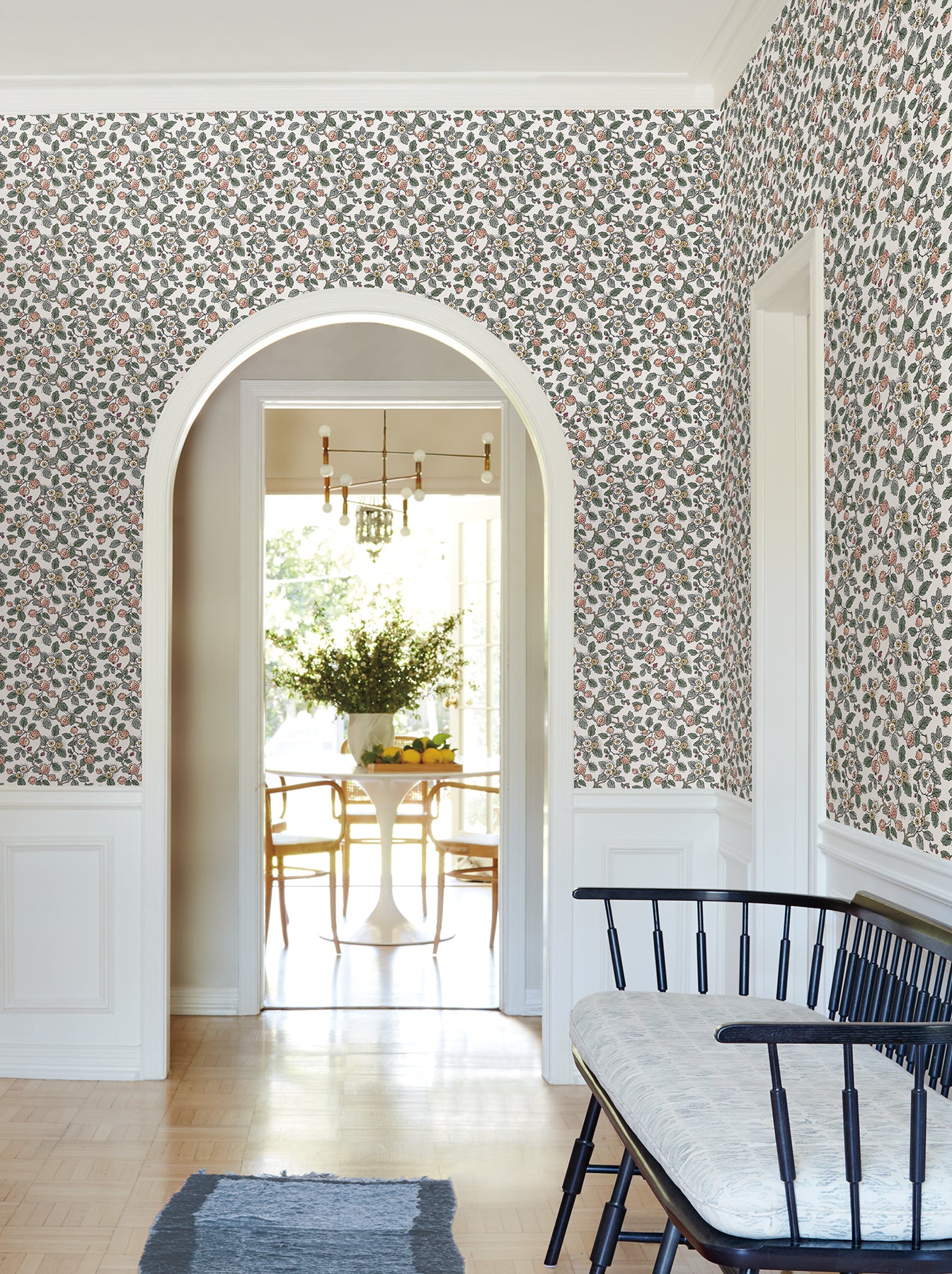 Arched entry to bright dining room with floral wallpaper, white wainscoting, black spindle bench, pale cushion, blue rug.