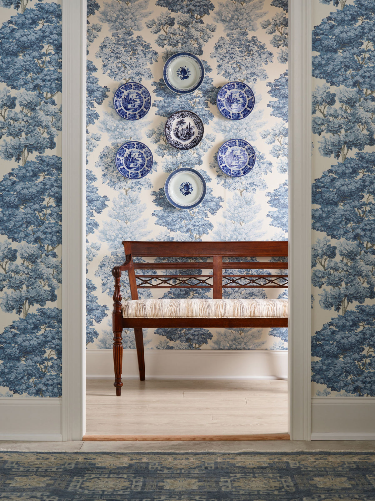 Brown wooden bench with carved backrest before blue-and-white toile wallpaper; wall plates in blue-and-white pattern above.