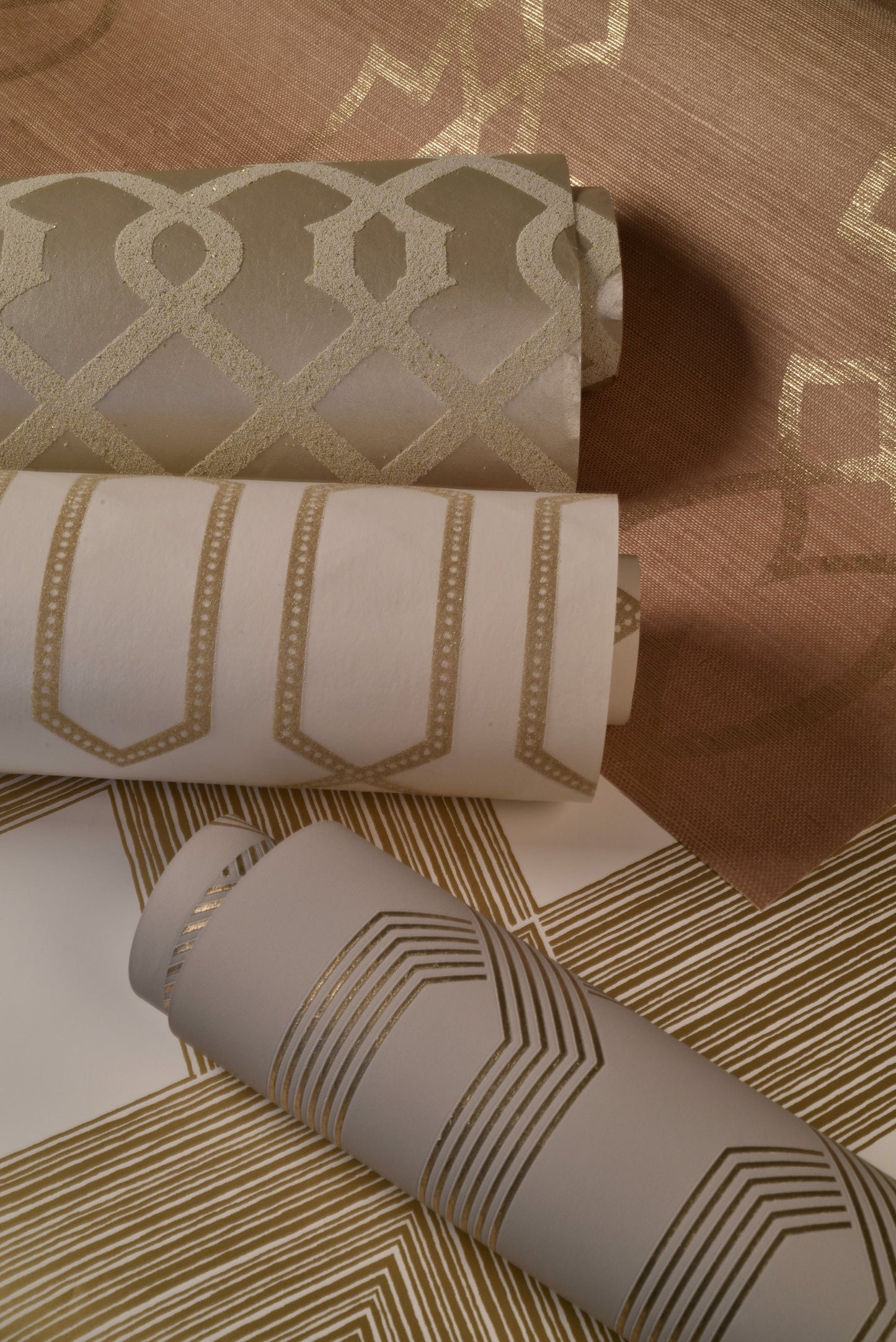 Three wallpaper rolls: beige-gold lattice, white roll with gold panel arches, and gray roll with gold chevrons.