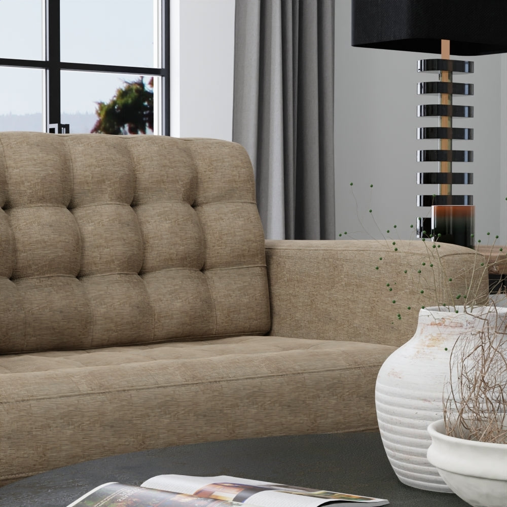 Beige tufted sofa upholstered in toast solid fabric with wide arms in a modern living room; gray curtains, striped lamp, and a white ribbed vase.