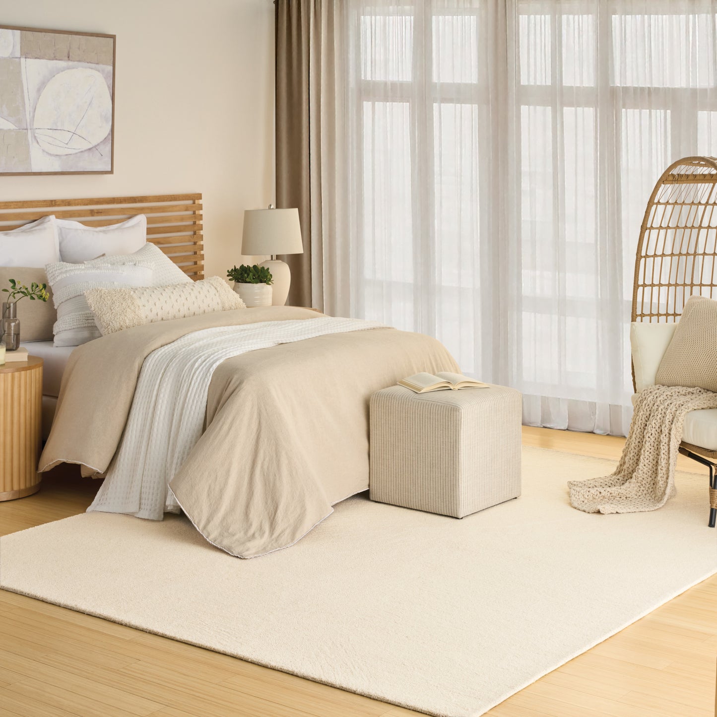 Neutral beige-gray rug on the floor in a room with a slatted wood bed, cream ottoman, wicker chair, and sheer curtains.