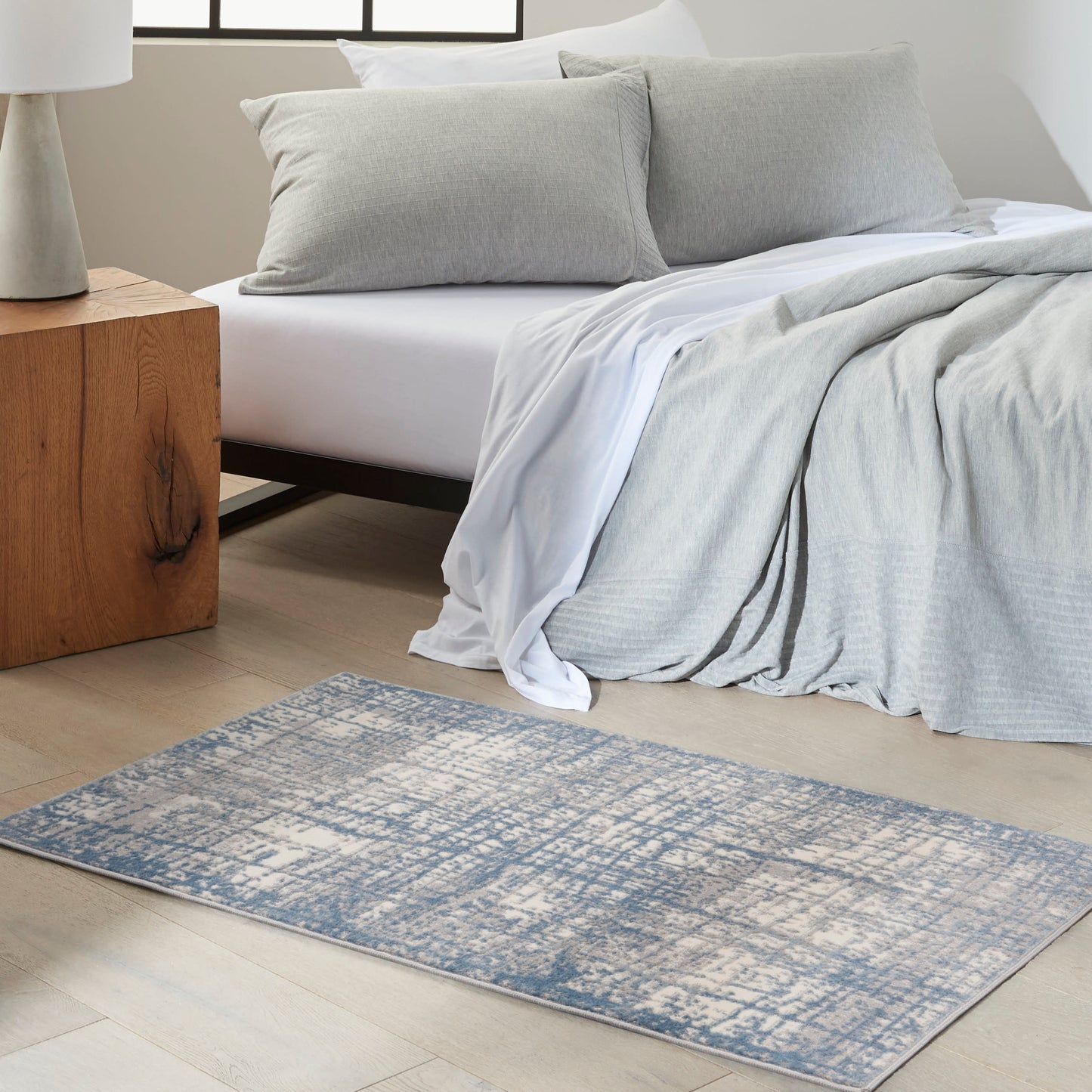 Rectangular blue-gray distressed area rug on light wood floor beside a white bed with gray duvet and pillows.