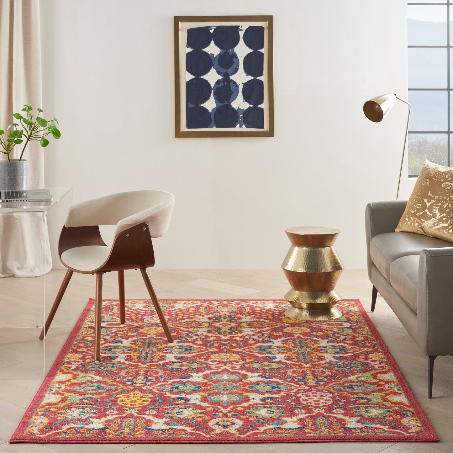 Vibrant rectangular area rug with red border and multicolor pattern on light wood floor, beside a mid-century chair and gray sofa.