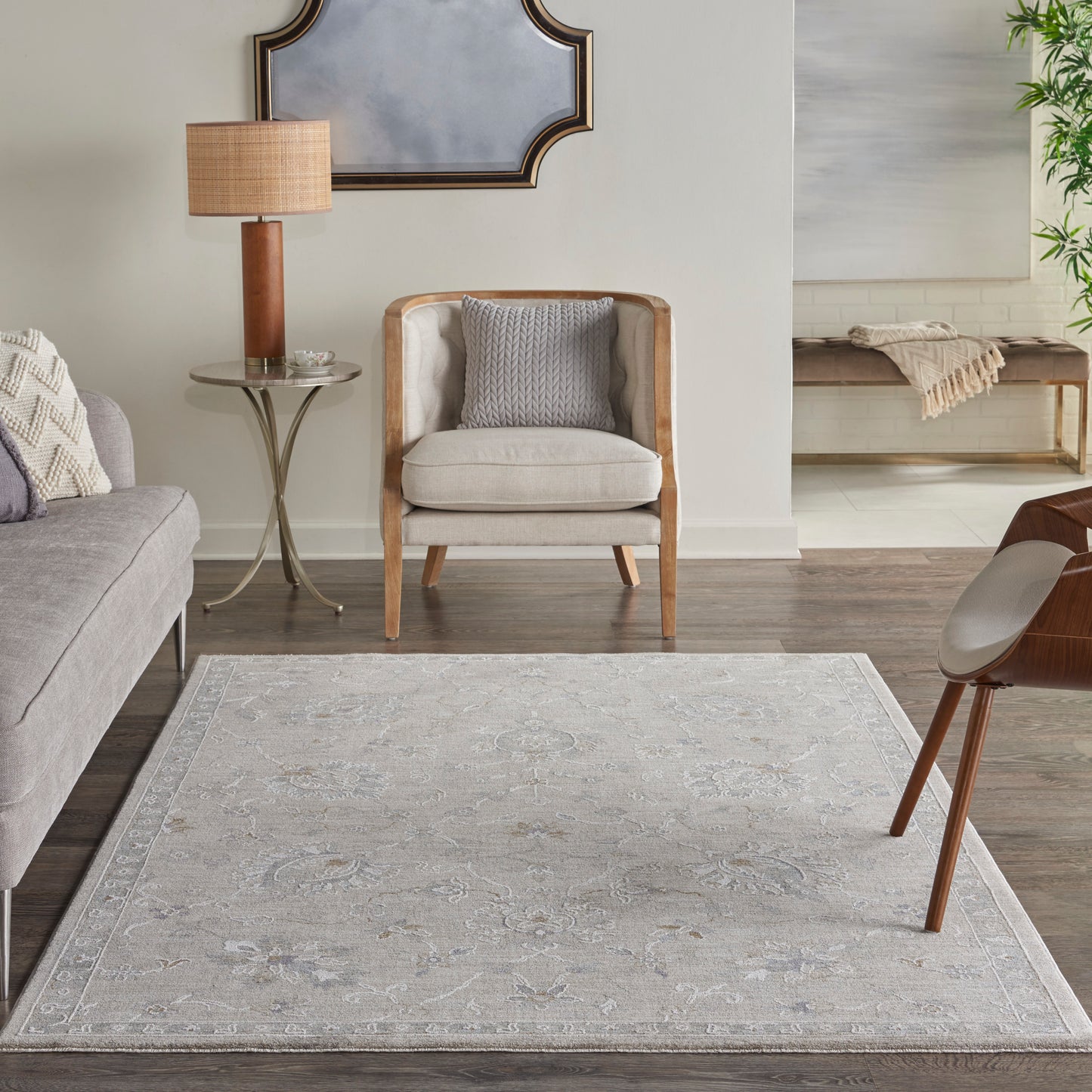 Neutral living room with gray fabric sofa, cream wood-frame chair, glass side table, wooden lamp, pale patterned rug.