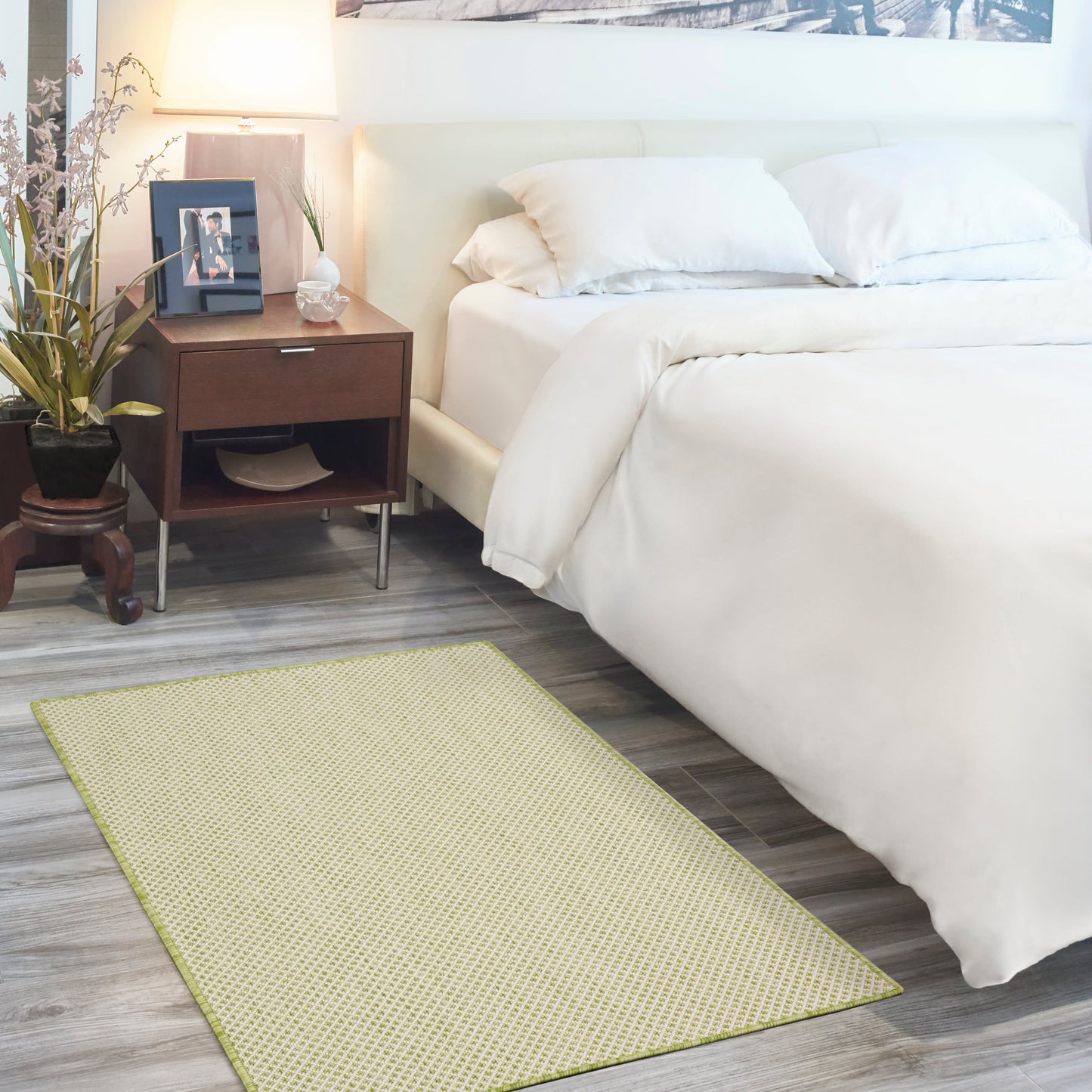 White bed with white duvet, dark wood nightstand, lamp, plant, and a yellow patterned rug on gray wood floor