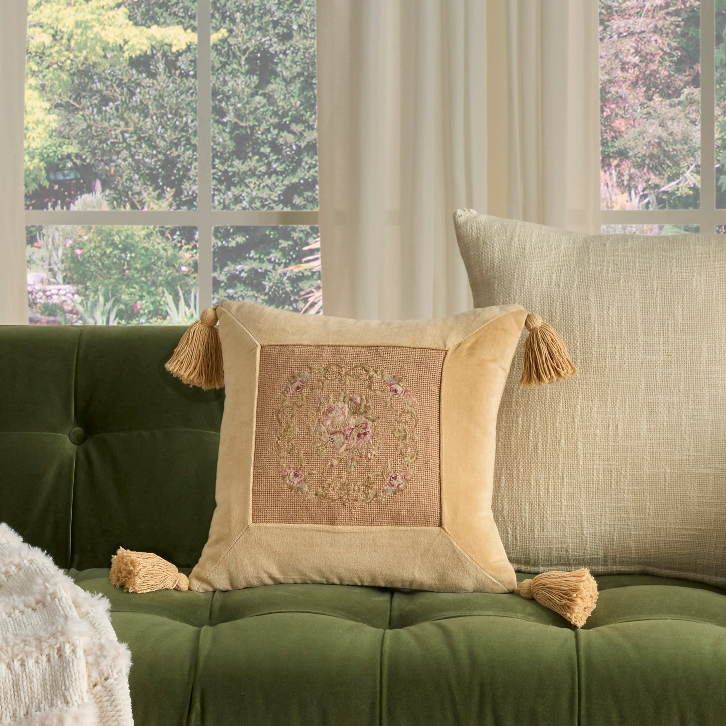 Blush decorative square throw pillow with tassels and embroidered center on a green tufted sofa in a sunlit living room