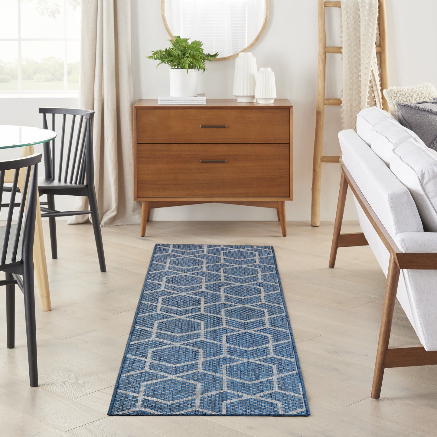 Blue geometric runner rug on light wood floor with a mid-century wooden dresser, sofa, and black chairs in a bright living room.