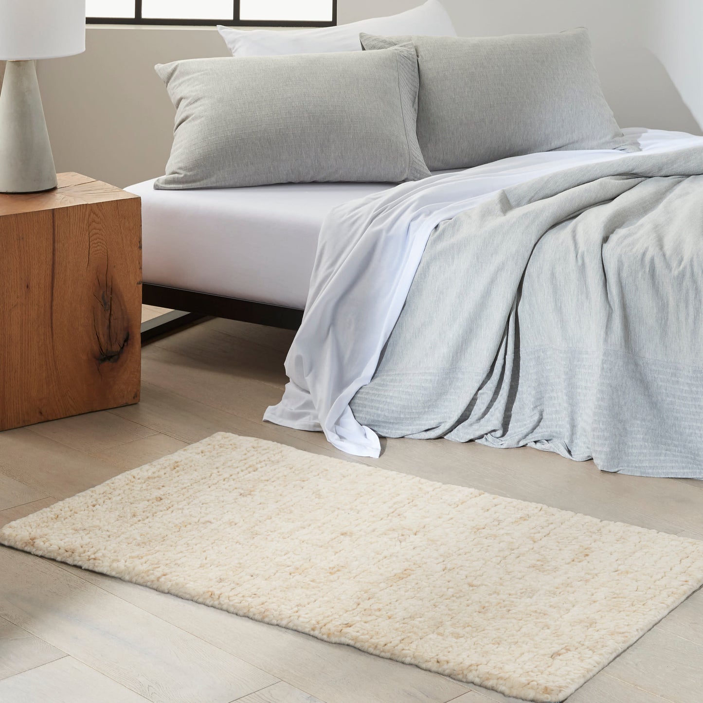 Cream shag rug in front of a white bed with light gray bedding and pillows, wood nightstand, airy modern bedroom.