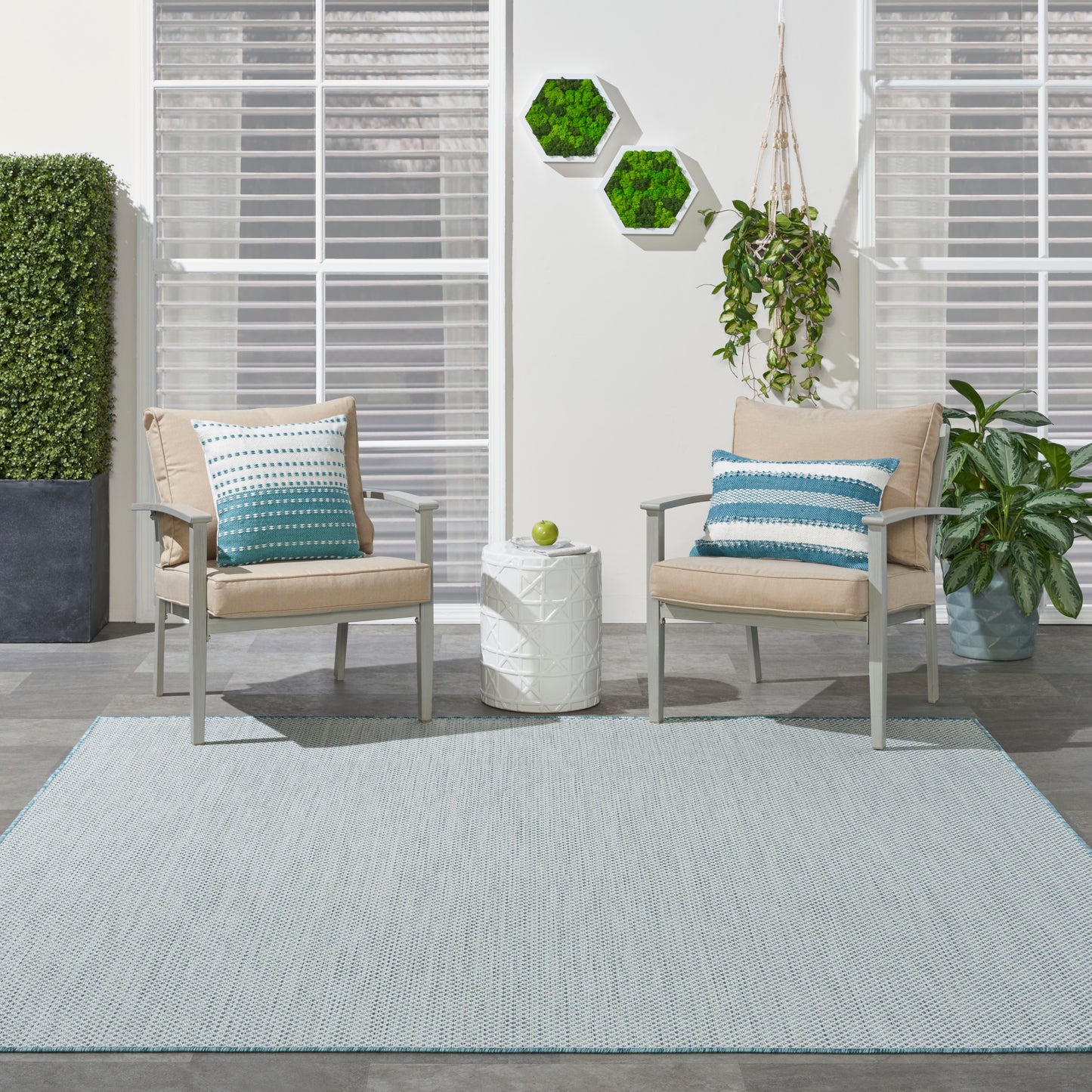 Light blue rug under two beige cushioned chairs on slender metal frames, turquoise striped pillows, a white textured side table, and abundant greenery.