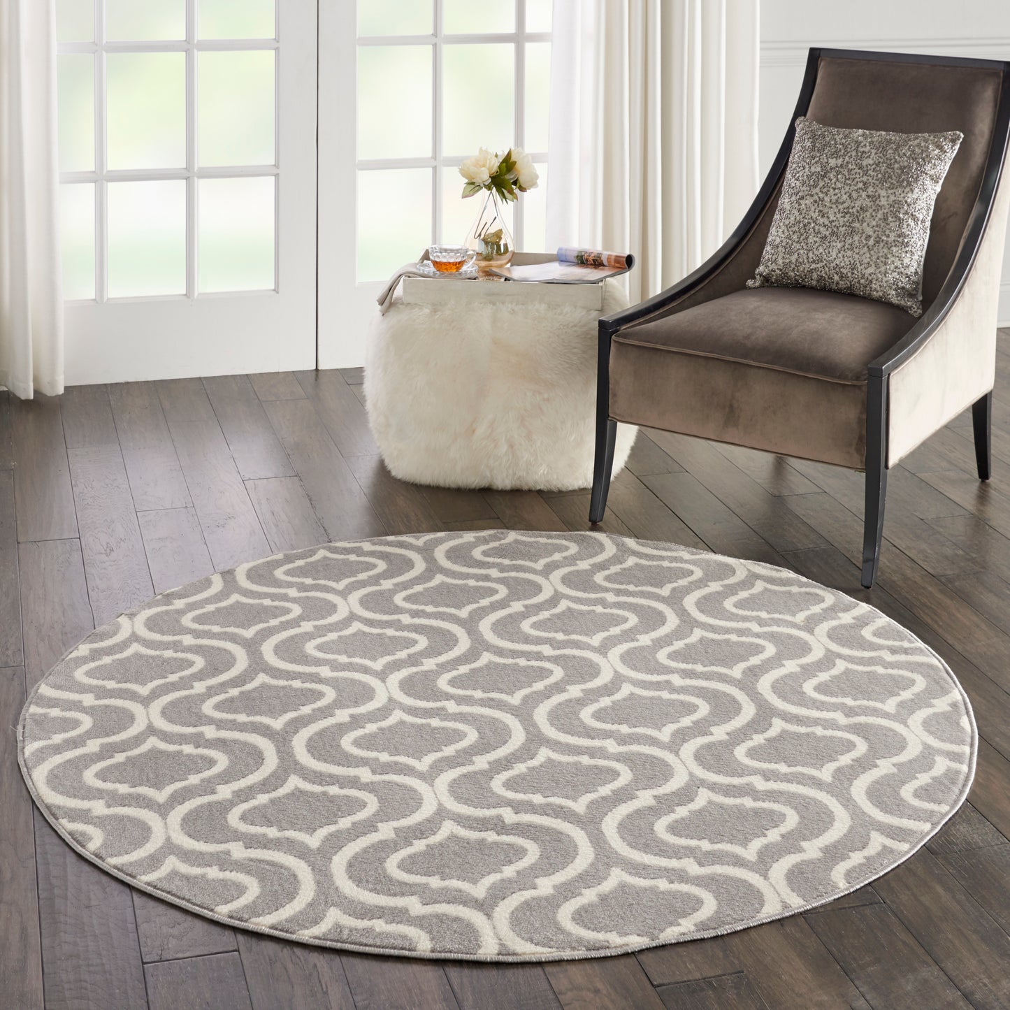 Round gray rug with white wavy lattice pattern on dark wood floor, beside a brown upholstered armchair and white fur ottoman.