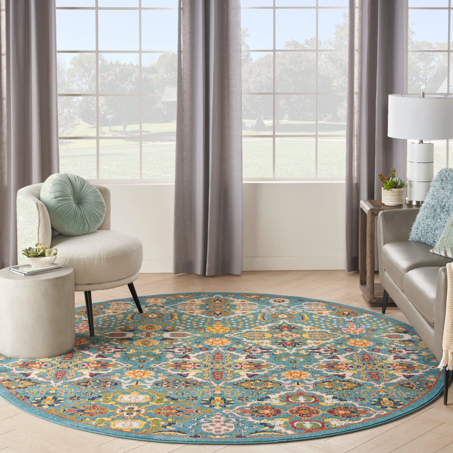Round teal rug with a colorful floral pattern in a cozy living room with a beige curved chair, gray curtains, and sofa.