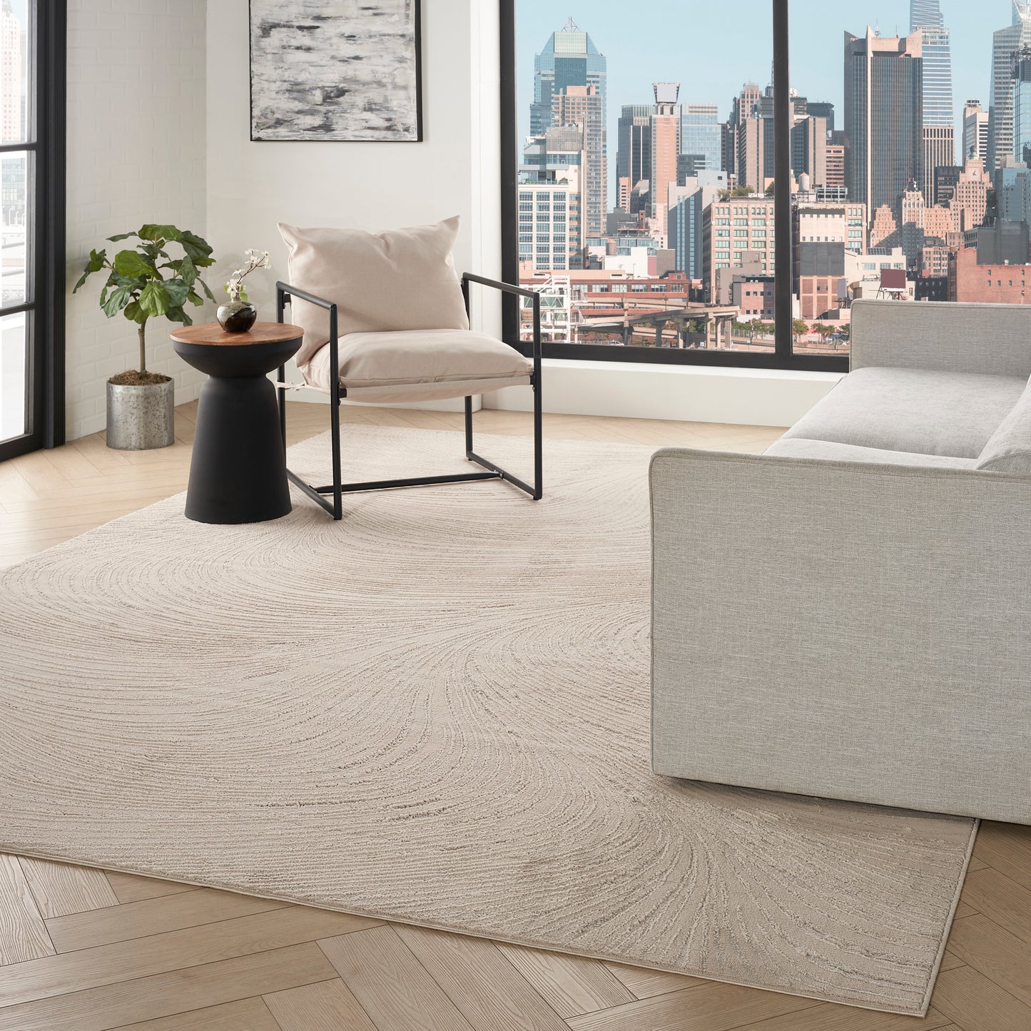 Neutral living room with city view window, beige rug on light wood floor, cream armchair, black side table, plant, gray sofa.