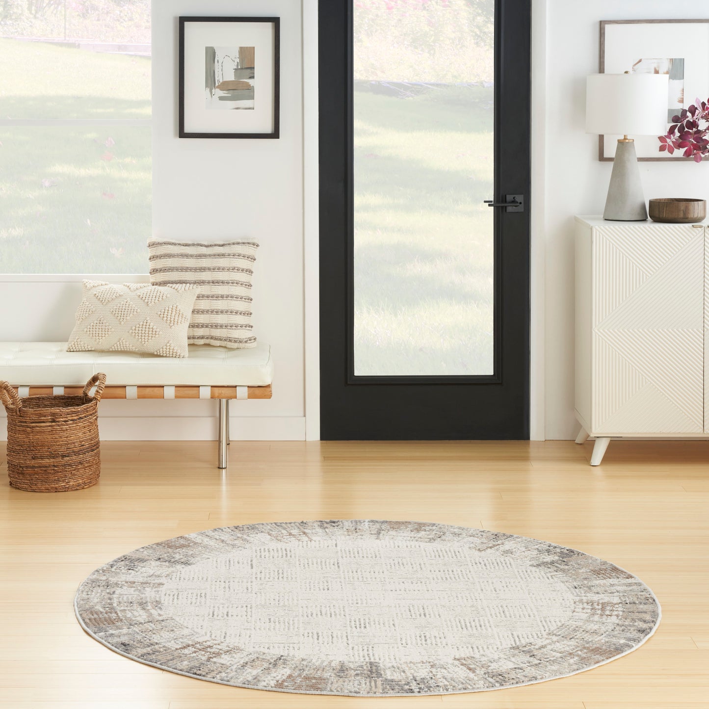Round gray textured rug on light wood floor in bright entry; bench with cushions, black-framed door, & white cabinet.