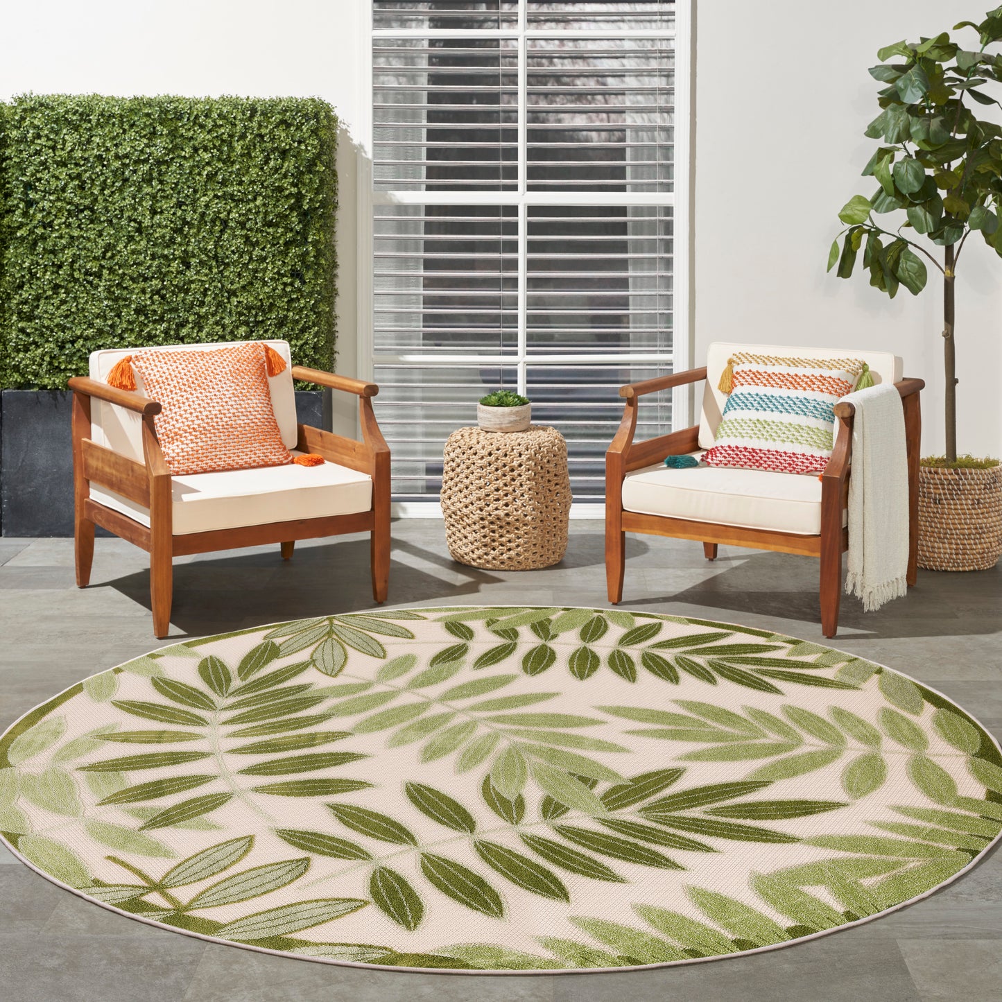 Round beige outdoor rug with green leaf pattern; wooden chairs with white cushions and colorful pillows, woven side table.