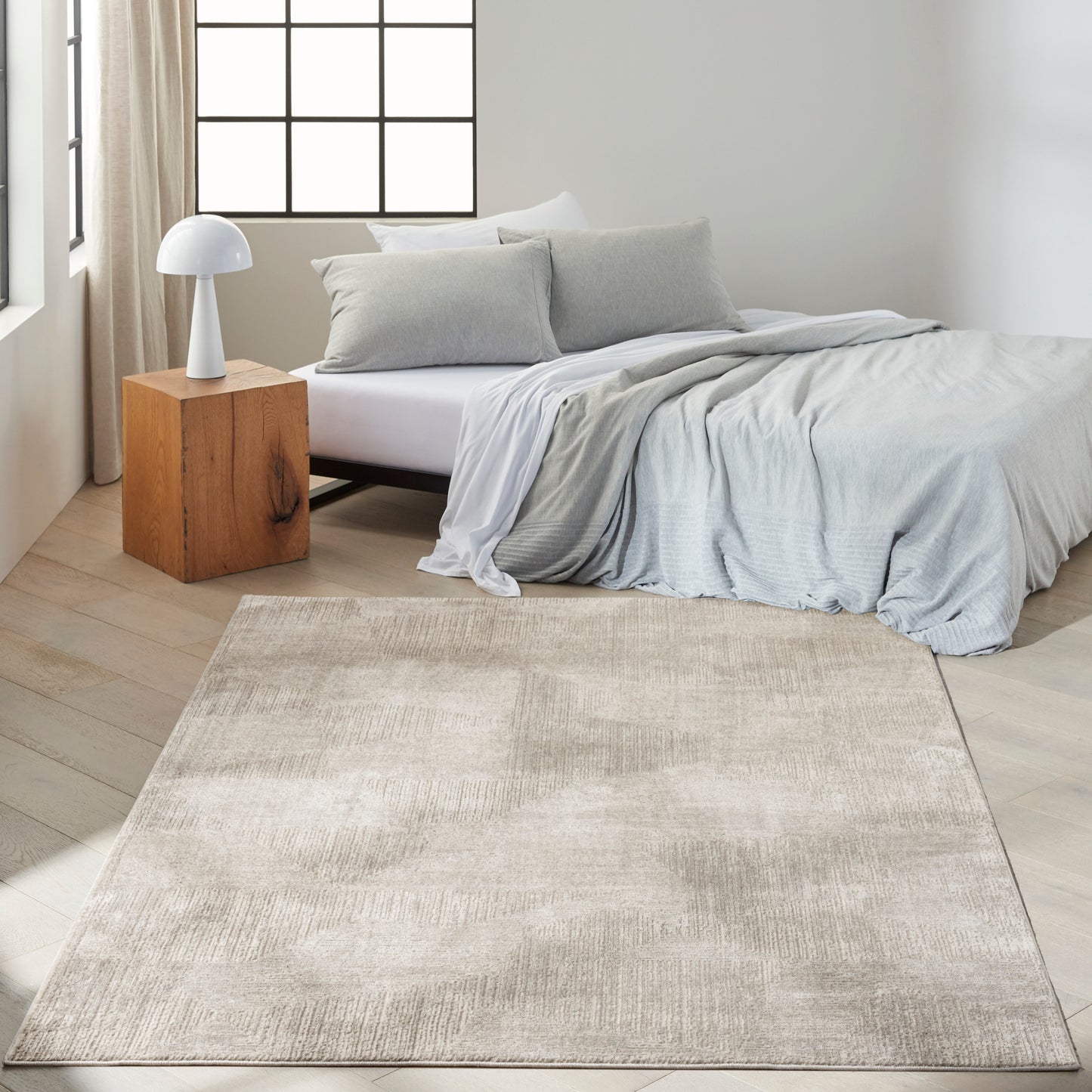 Modern bedroom with light gray bedding on a low bed, beige area rug, wooden cube nightstand and white mushroom lamp.