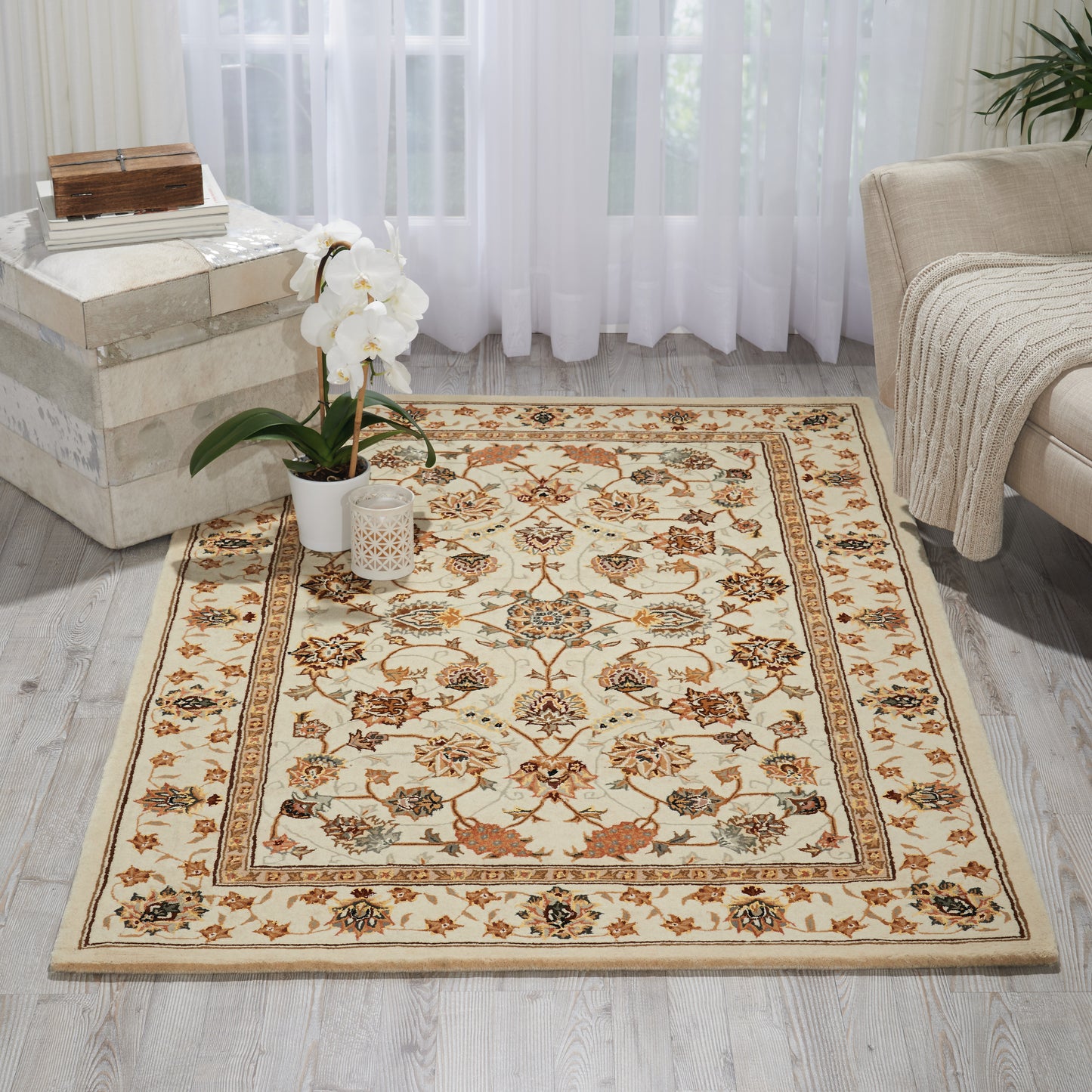 Cream floral area rug with ornate beige border, orange accents, on light wood floor beside a potted orchid and beige sofa.