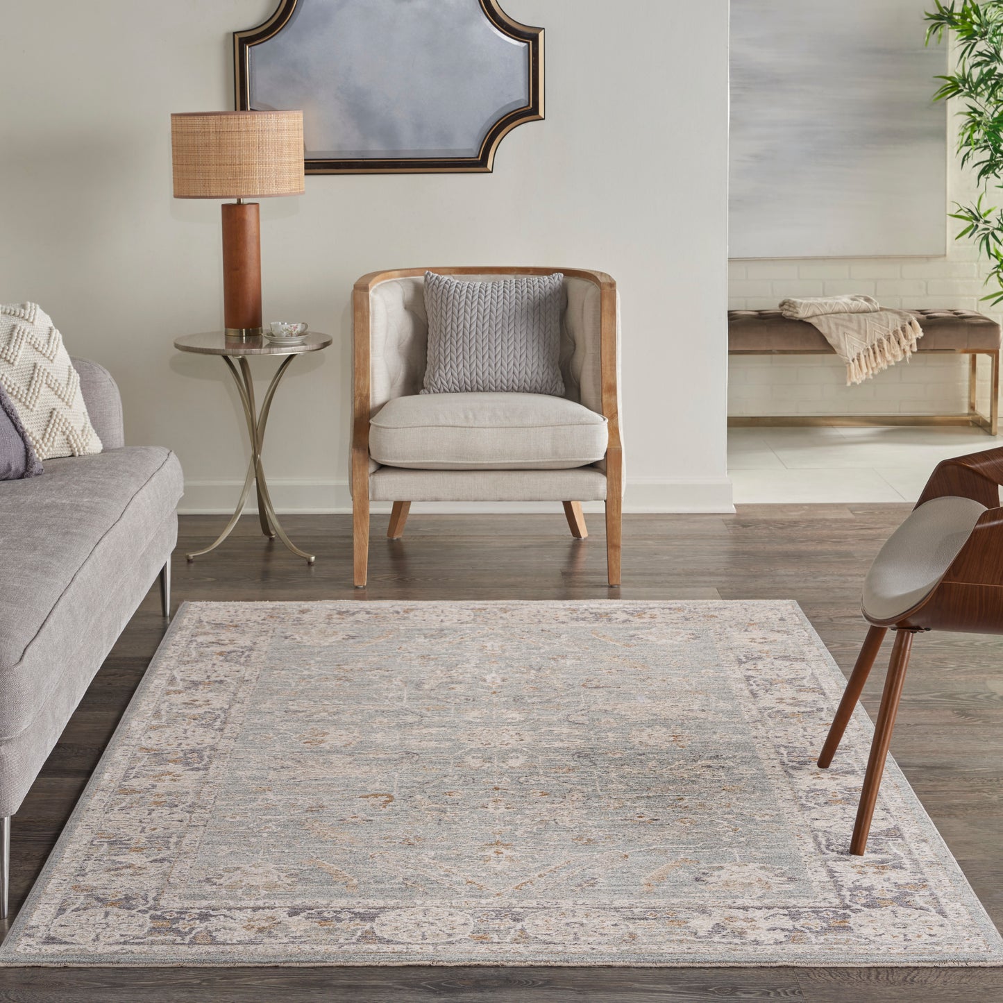 Neutral living room with pale ornate-border rug, light wood armchair, gray cushions, gray sofa, brass round side table.