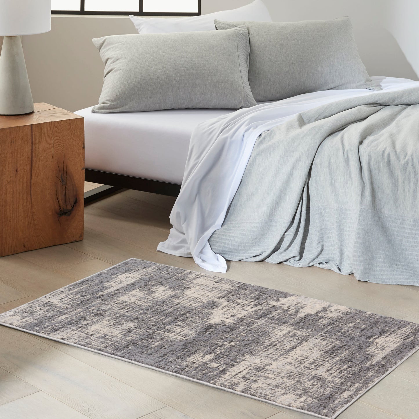 Minimalist bedroom: light gray duvet and pillows, draped gray throw, concrete lamp on wooden nightstand, textured gray rug.