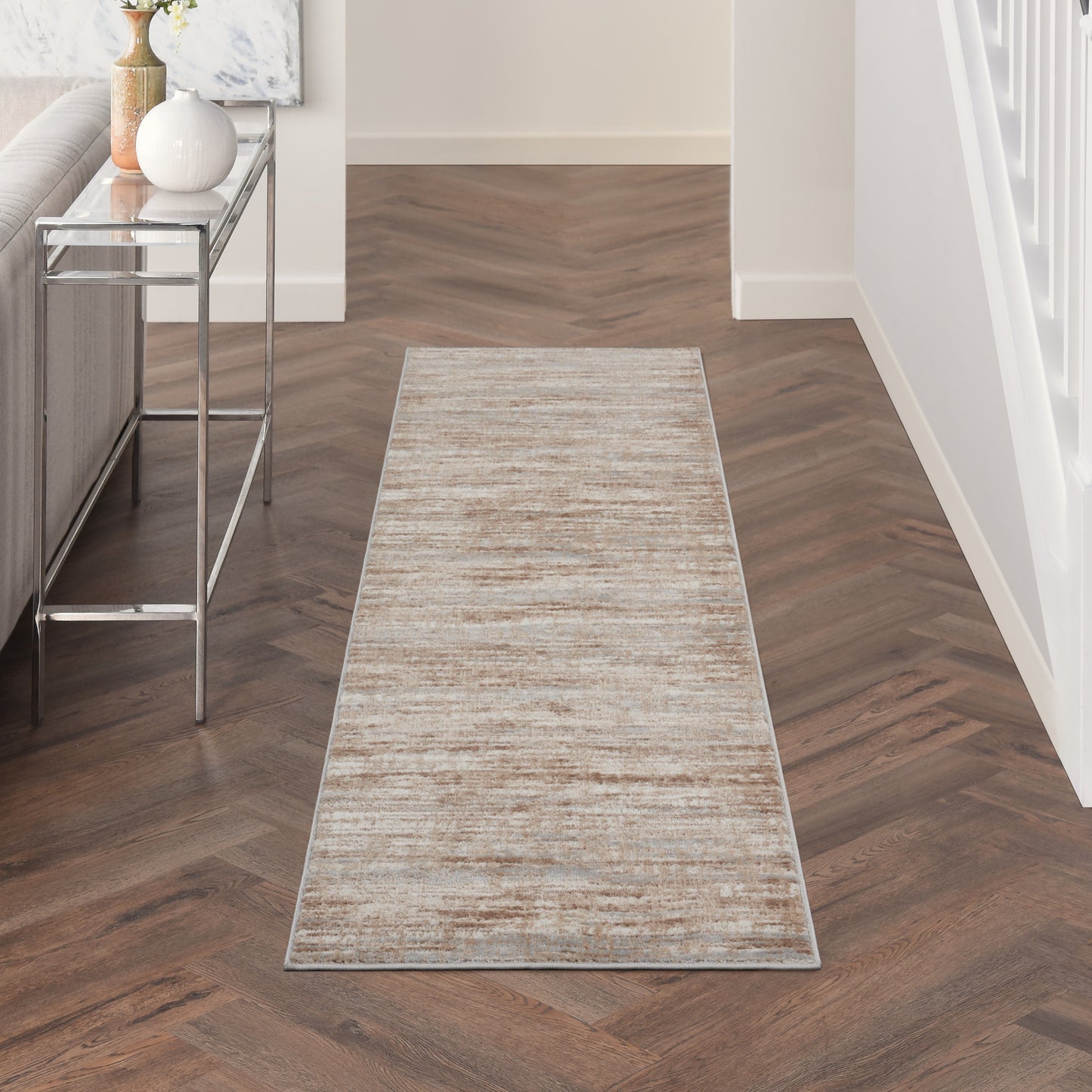 Beige textured hallway runner rug on dark herringbone wood floor; chrome console table with vase to the left.