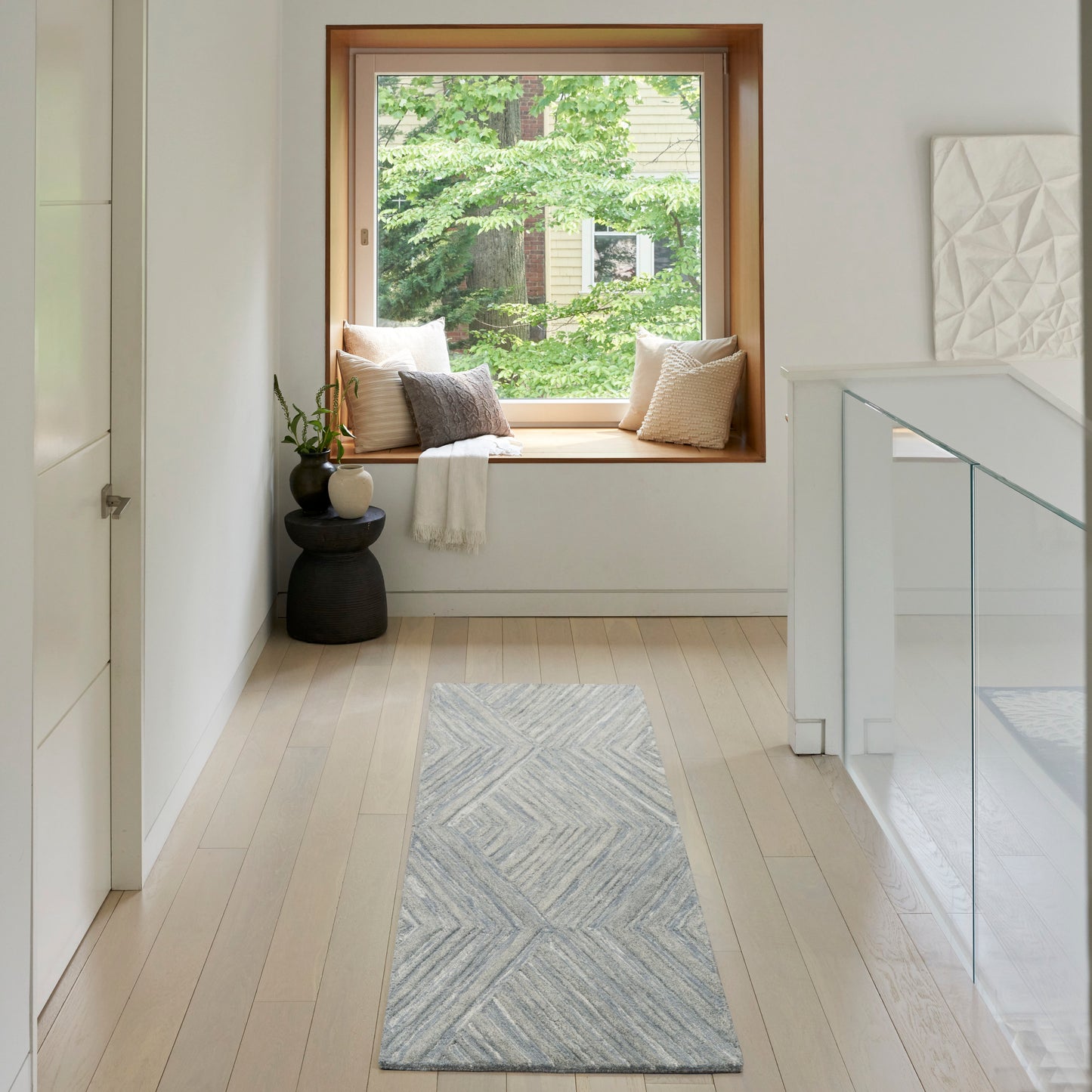 Bright hallway with pale wood floors, built-in window seat with cushions, blue-gray chevron runner rug, greenery outside window.
