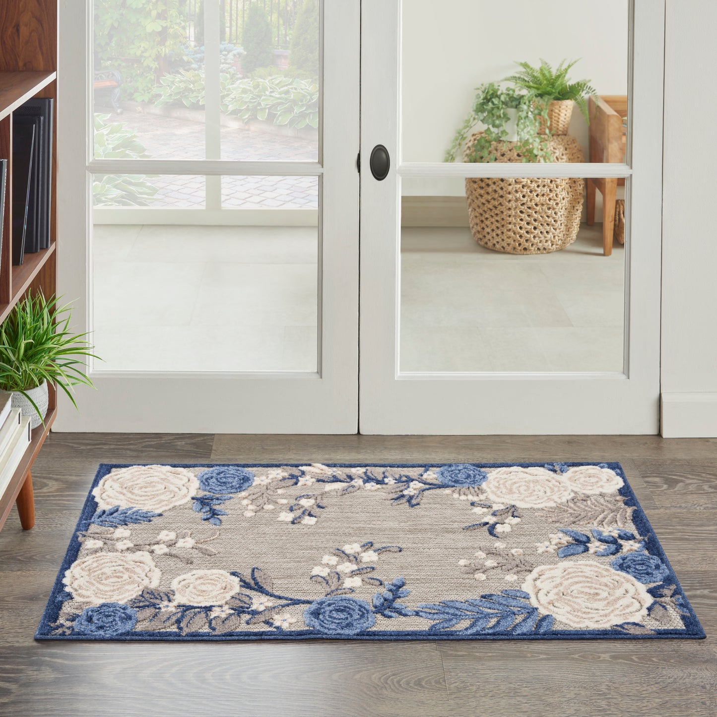 Blue floral outdoor rug with white and cream flowers on a gray-beige field and dark blue border, laid on a wooden deck by glass doors.