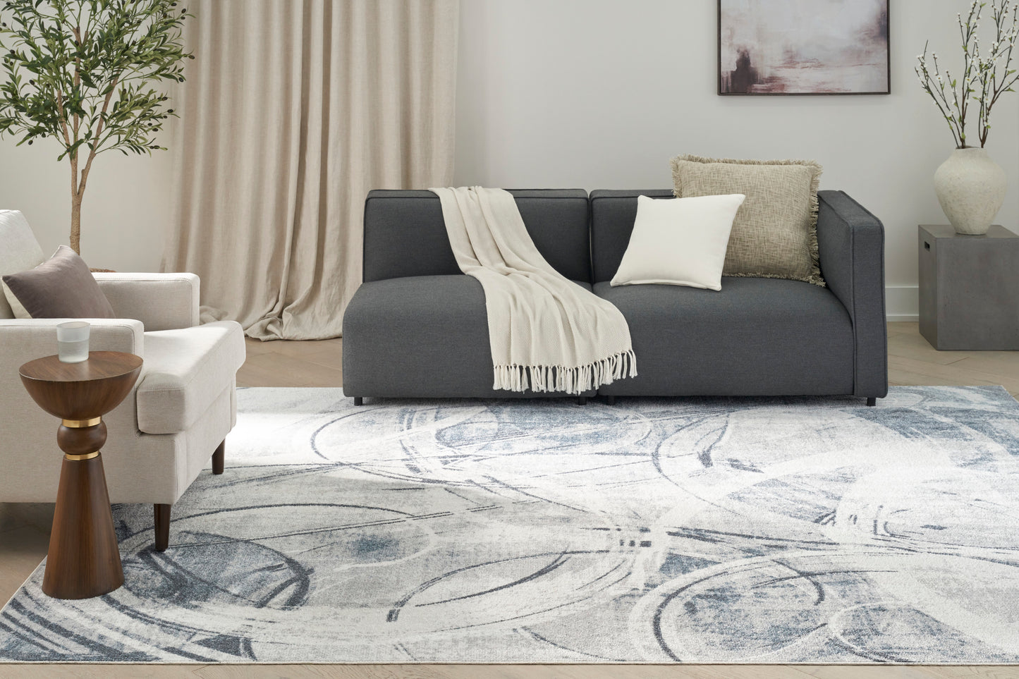 Modern living room with charcoal gray sofa, cream throw, white armchair, wood side table, blue-gray patterned rug.