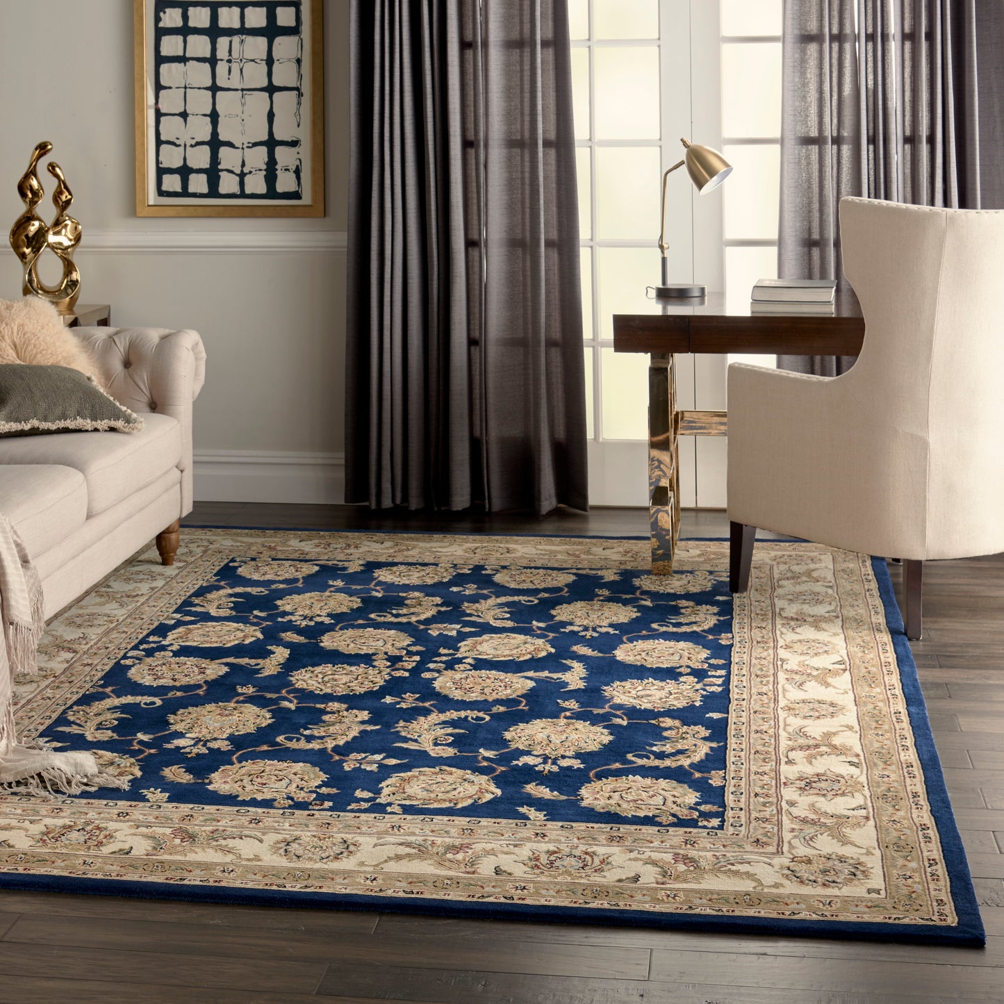 Blue ornate rug with beige floral pattern & border in a modern living room with cream sofa, beige armchair, & desk.