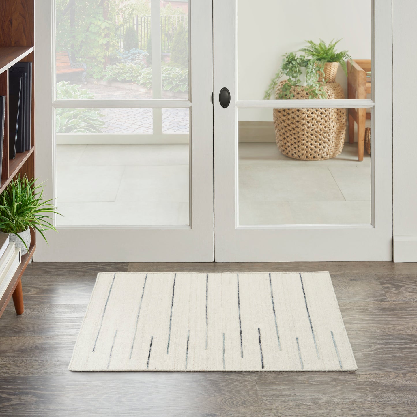 White French doors with glass panels open to a sunny garden; cream rug with blue stripes on dark wood floor, potted plants.