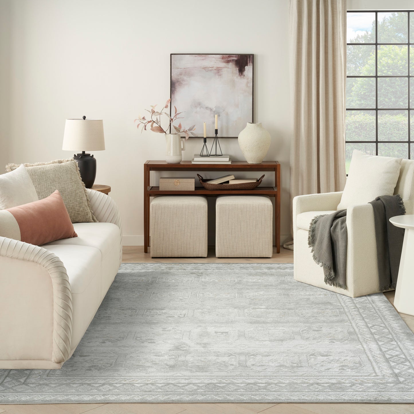 Neutral living room with cream sofa, beige armchair, light gray patterned rug, wooden console, ottomans, vase, abstract art.