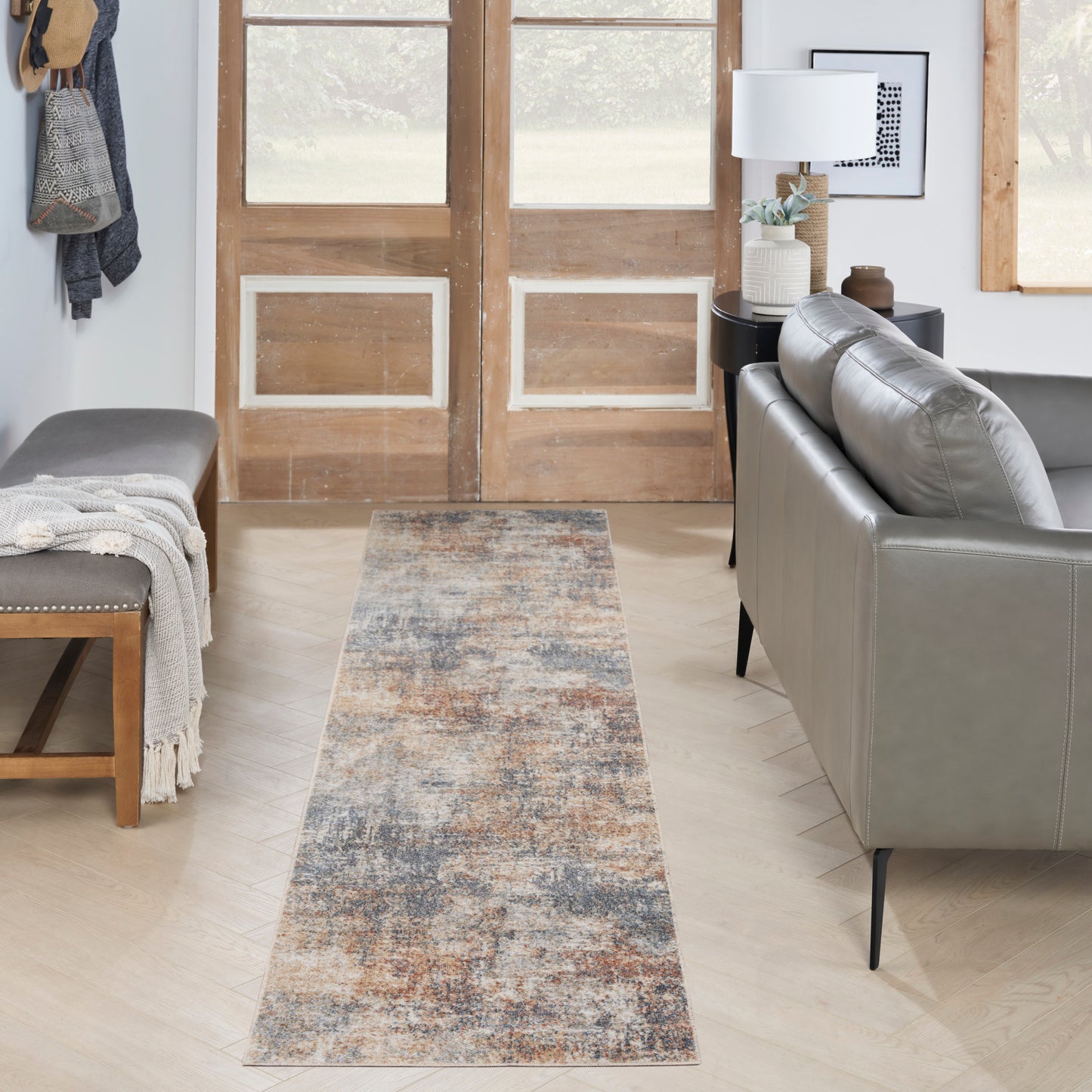 Long abstract rug runner in blue, brown and gray on a light wood floor, flanked by a gray sofa and wooden entry doors.