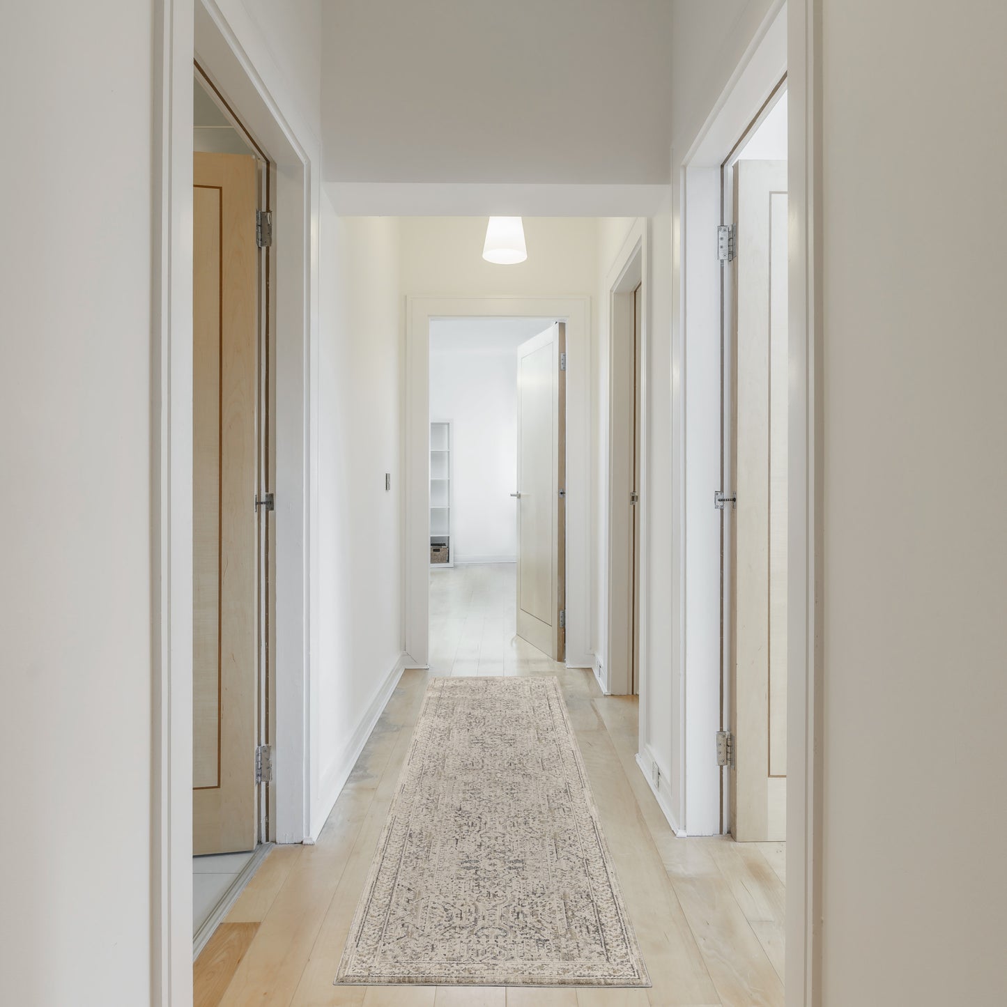 Bright hallway with light wood doors, white walls, beige patterned runner rug, and a pendant light at the far end.