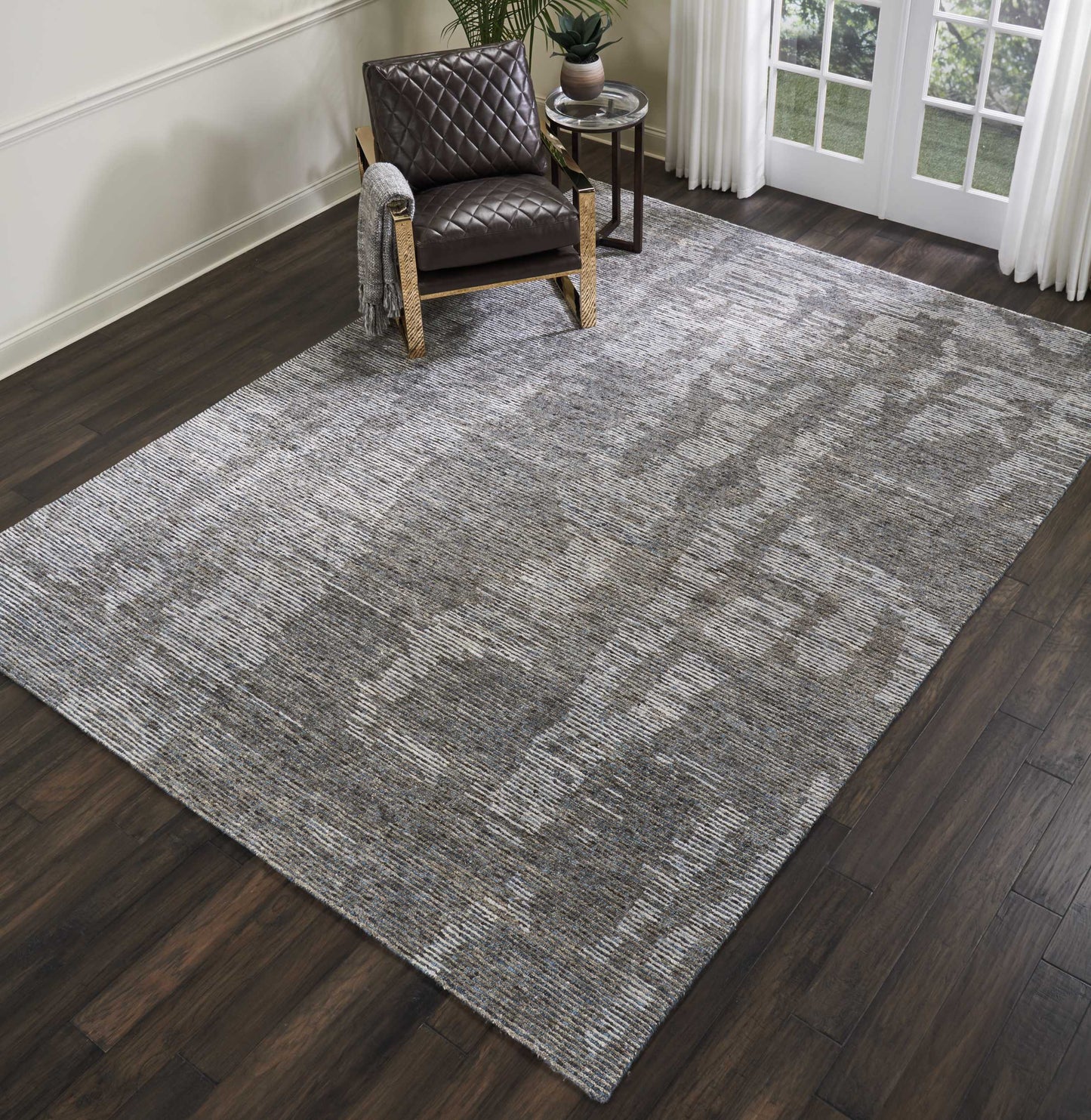 Large gray textured area rug on dark wood floor with a brown quilted armchair, a small side table, plant, and curtains.