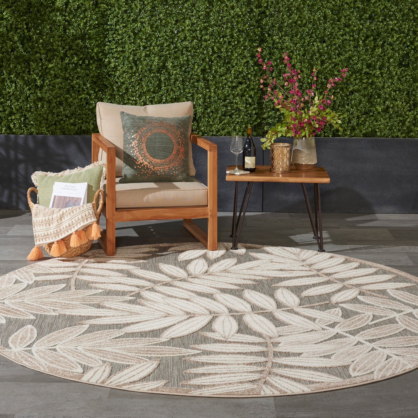 Outdoor seating area with round beige leaf-pattern rug, wooden armchair, small table with wine, vase of pink flowers.