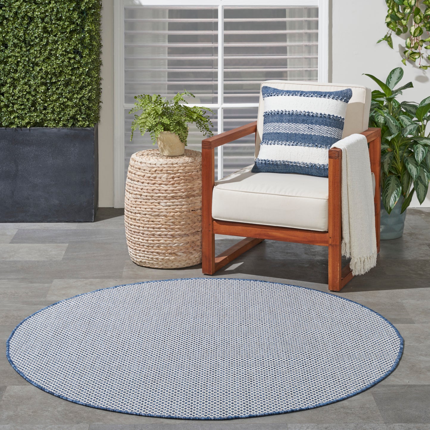 Round blue-and-white outdoor rug with wooden armchair, cream cushions, striped pillow, and woven side table with plant.