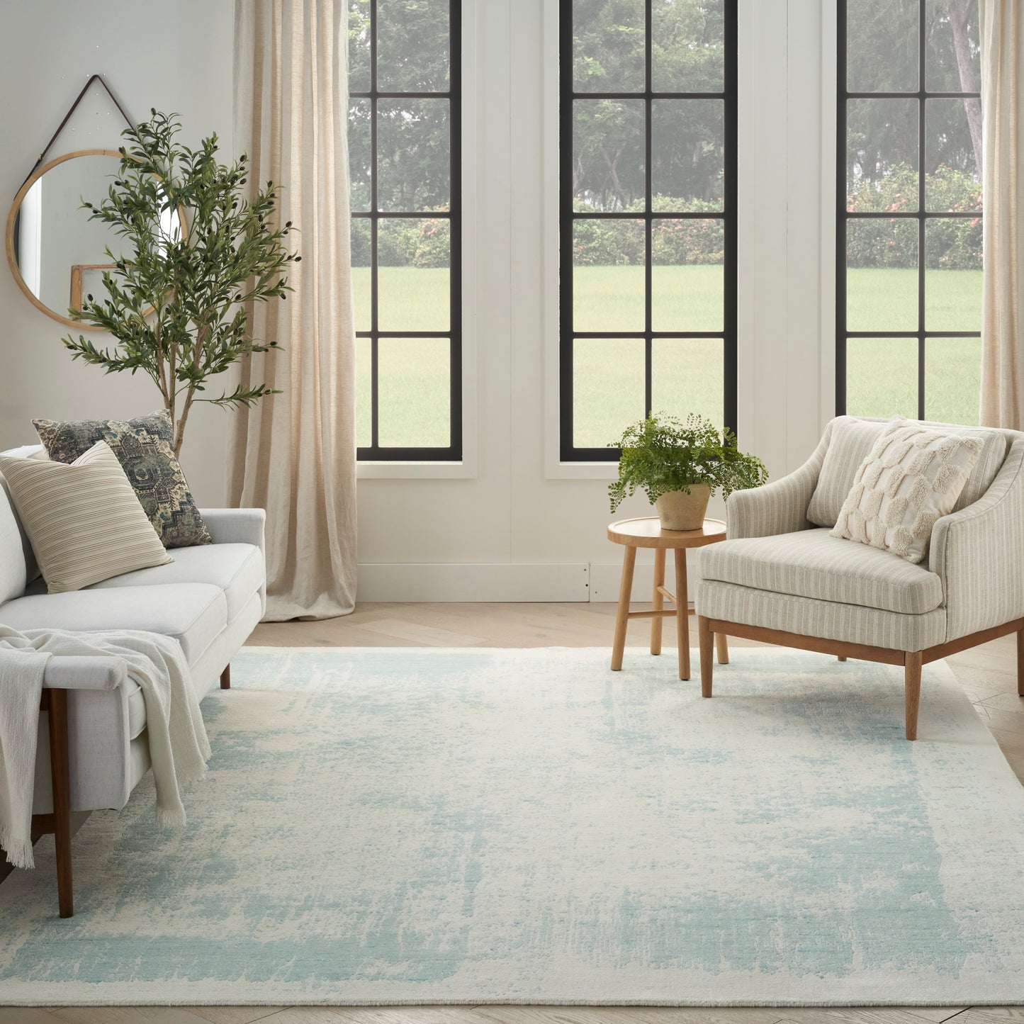 Bright living room with cream sofa, beige armchair, light wood legs, blue rug, potted plant, round mirror, grid windows.