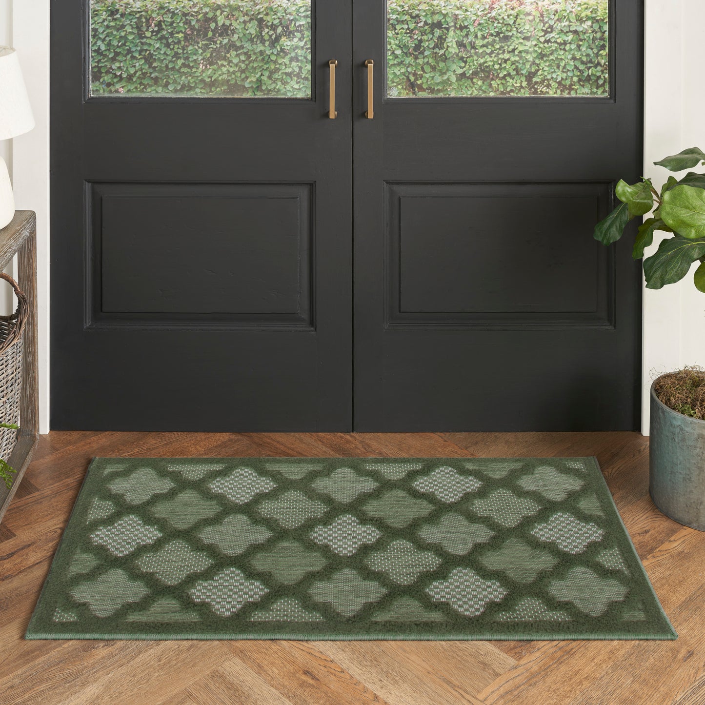 Green lattice-pattern rug on wood floor in front of double black doors with brass handles; potted plant nearby.