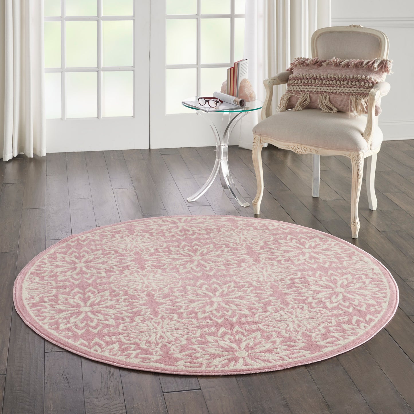 Pink round floral area rug on dark wood floor, with a vintage cream chair and glass side table in a bright living room.
