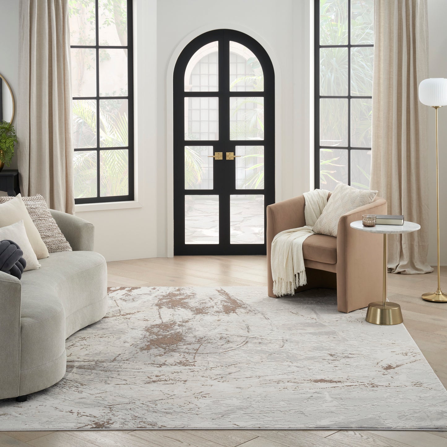 Bright living room with arched black door, beige curtains, light gray sofa, tan armchair, gold floor lamp, round side table & pale rug.