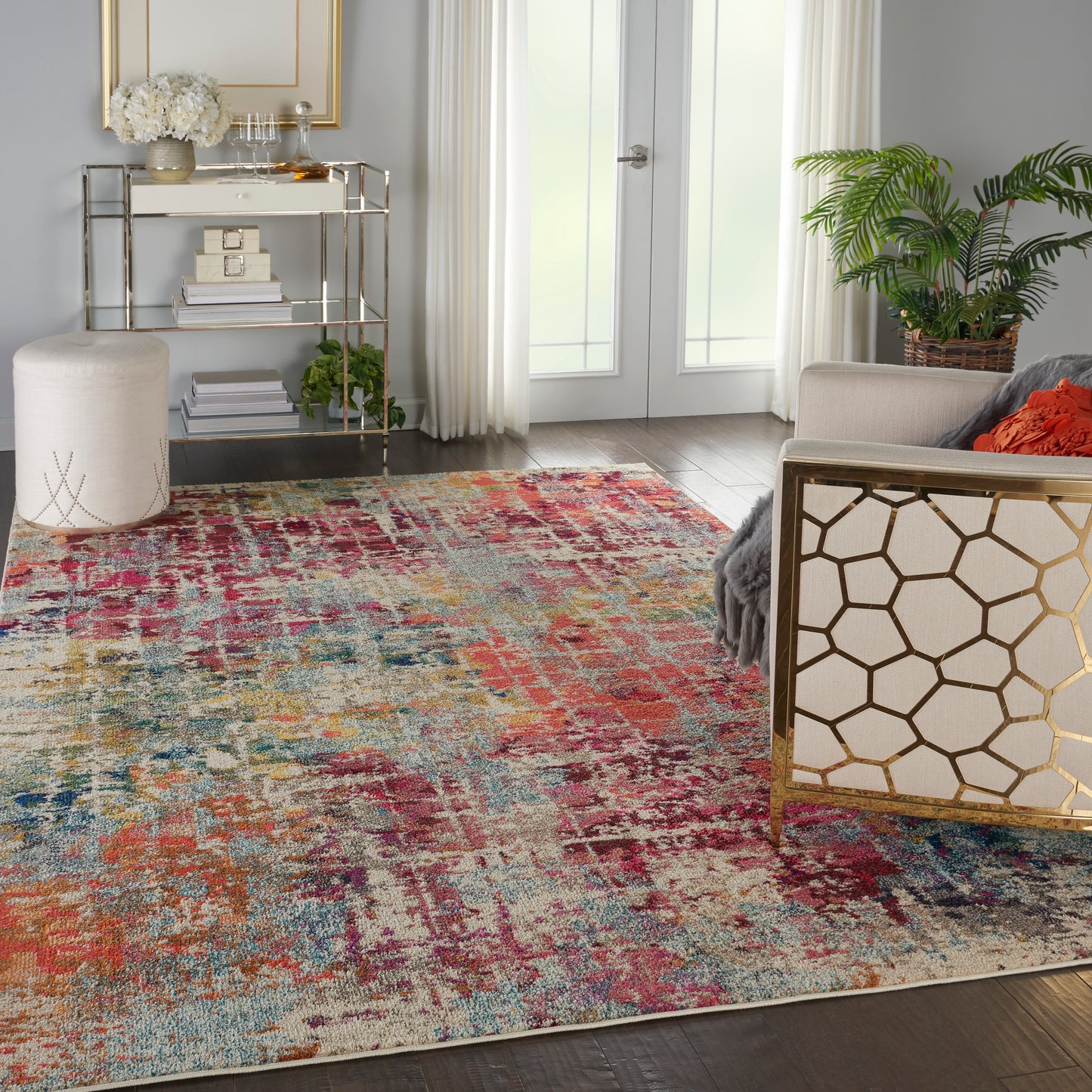 Colorful abstract area rug in a bright living room with gold lattice chair, glass bookshelf, potted plant, and cream seating.