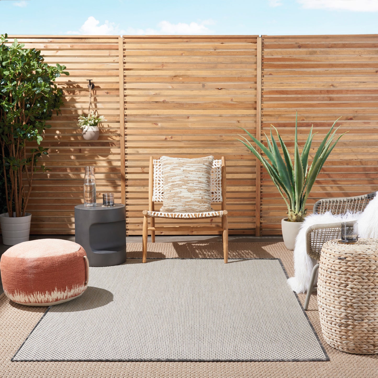 Sunny outdoor patio with natural wood fence, wicker chair with beige cushion, coral pouf, potted plants, and woven rug.