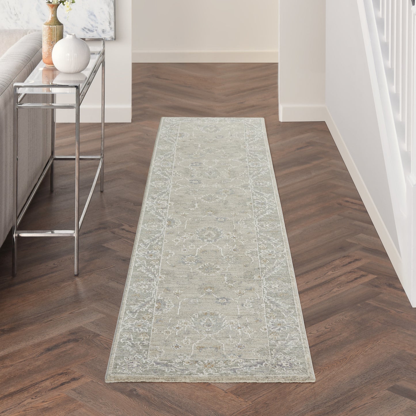 Vintage 10' runner rug in light gray with an ornate border, in a wood herringbone hallway; modern glass-topped console on the left.