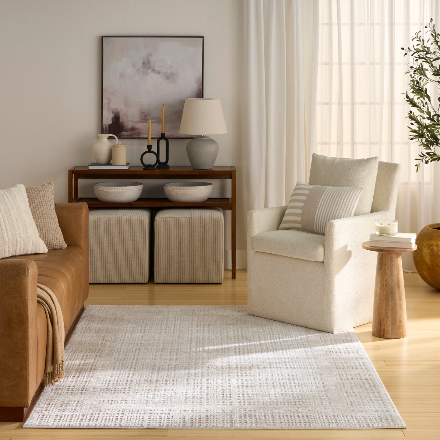 Cozy neutral living room: tan leather sofa, cream armchair, light beige rug, wooden console with decor, two ottomans.