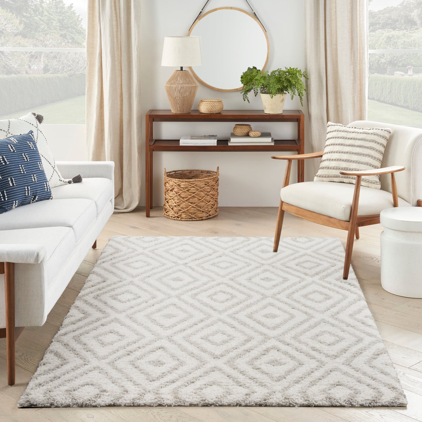 Neutral living room with cream sofa, mid-century chair, round mirror, wooden console, wicker basket, and diamond-pattern rug.