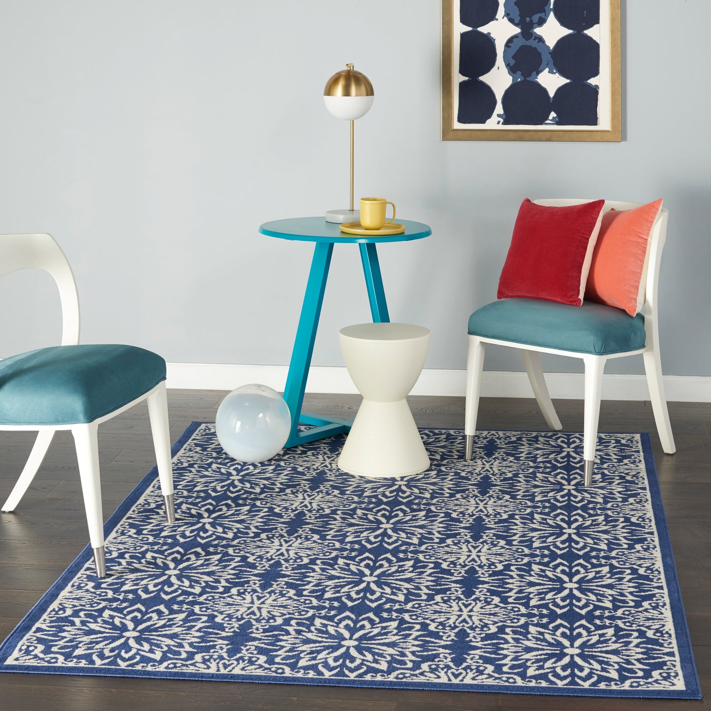 Teal round table with white/teal chairs, red and orange cushions, blue floral rug, gold lamp, and abstract wall art.