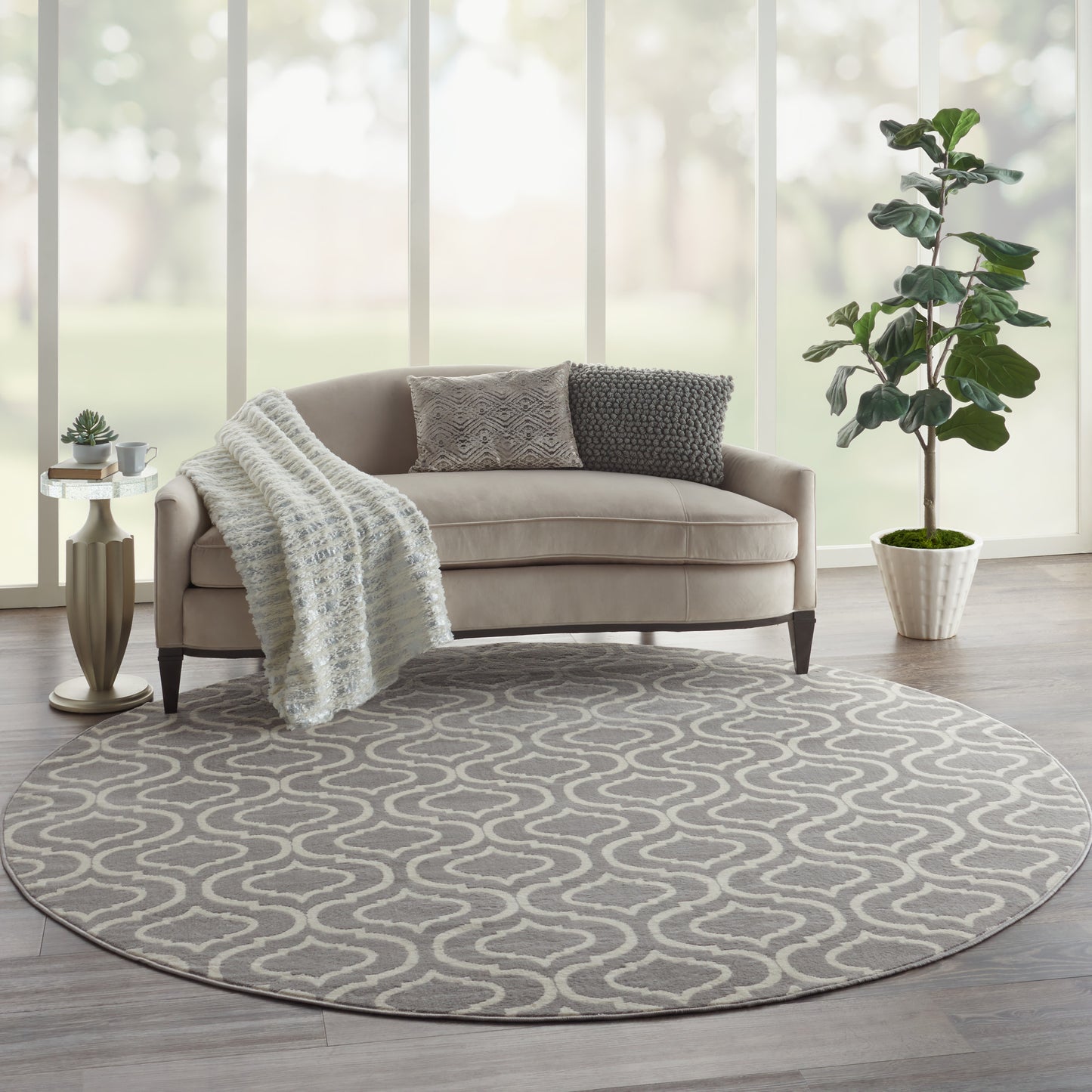 Round grey area rug with white abstract loop pattern in a modern living room; beige sofa, knitted throw, and potted plant.