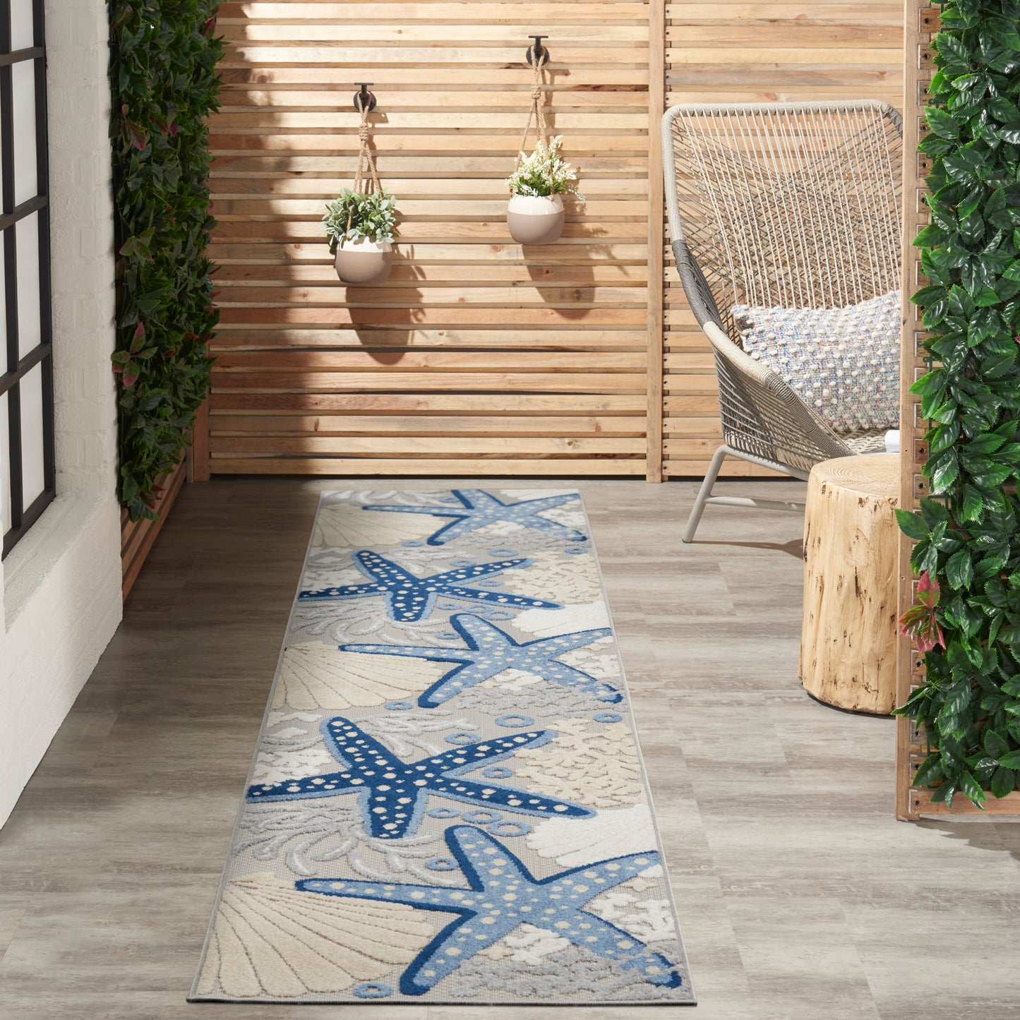 Coastal blue starfish runner rug on a light wooden patio with wicker chair & hanging planters.