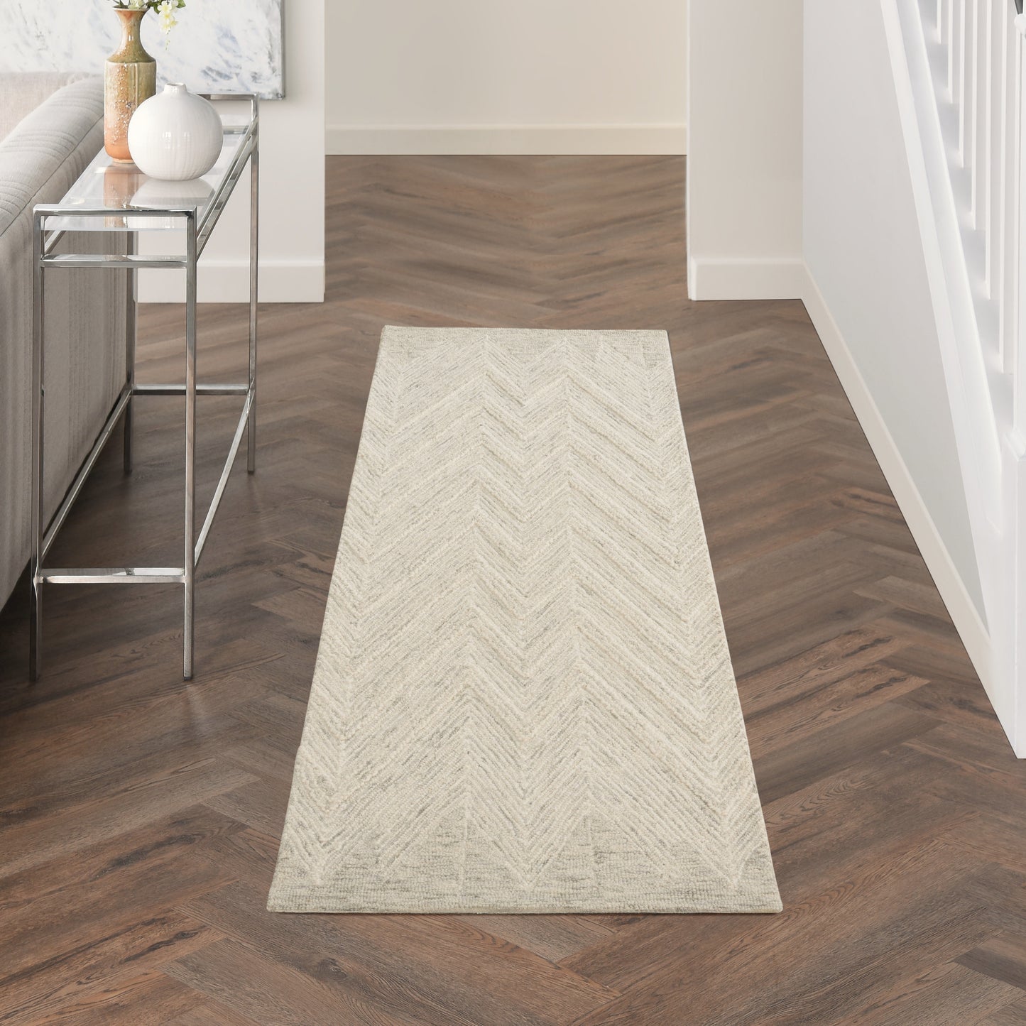 Beige chevron-patterned runner rug in a hallway with dark wood herringbone floor and a glass console table.