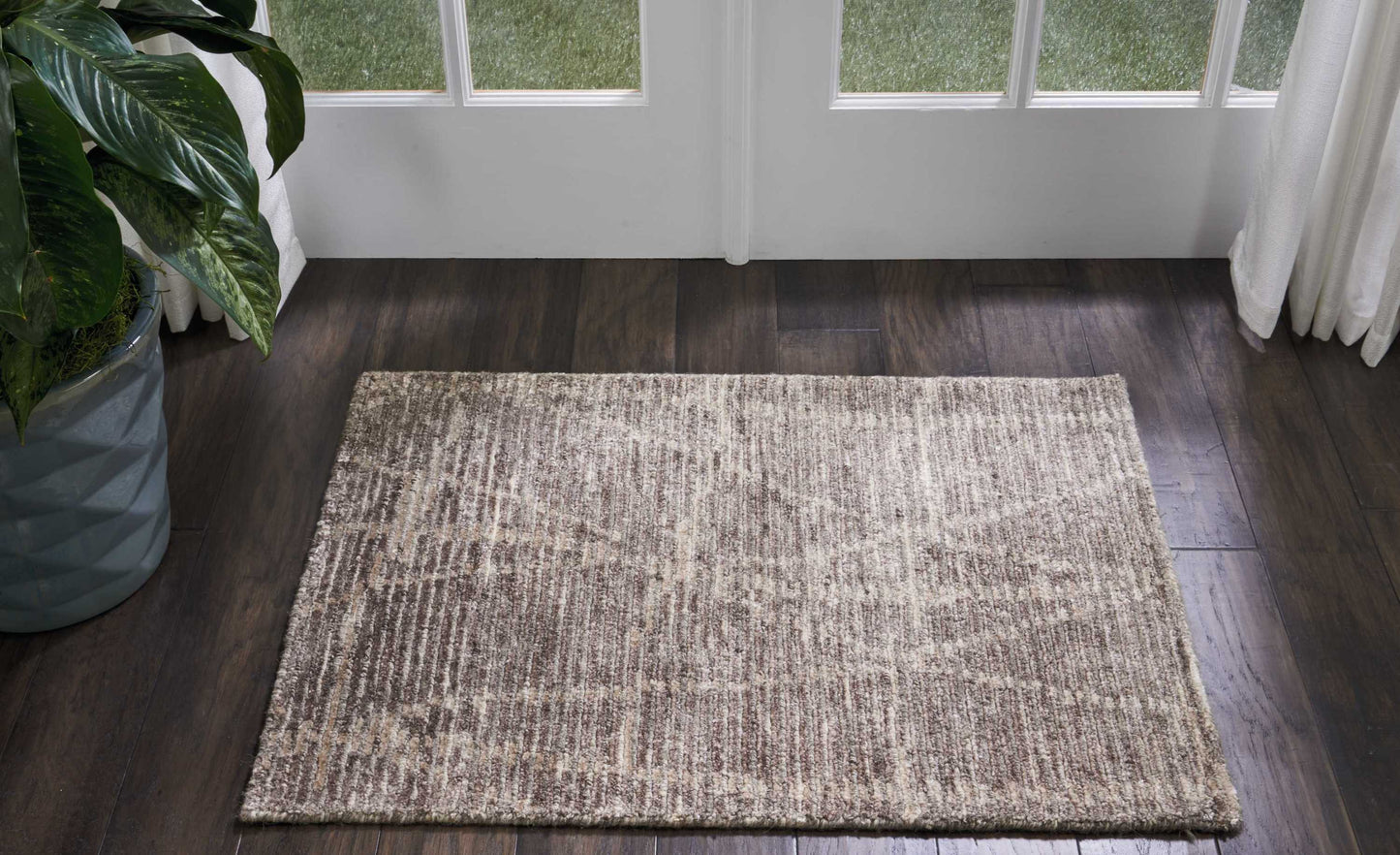 Neutral beige-brown rectangular rug with a subtle mottled pattern on a dark wood floor beside glass doors and a potted plant.