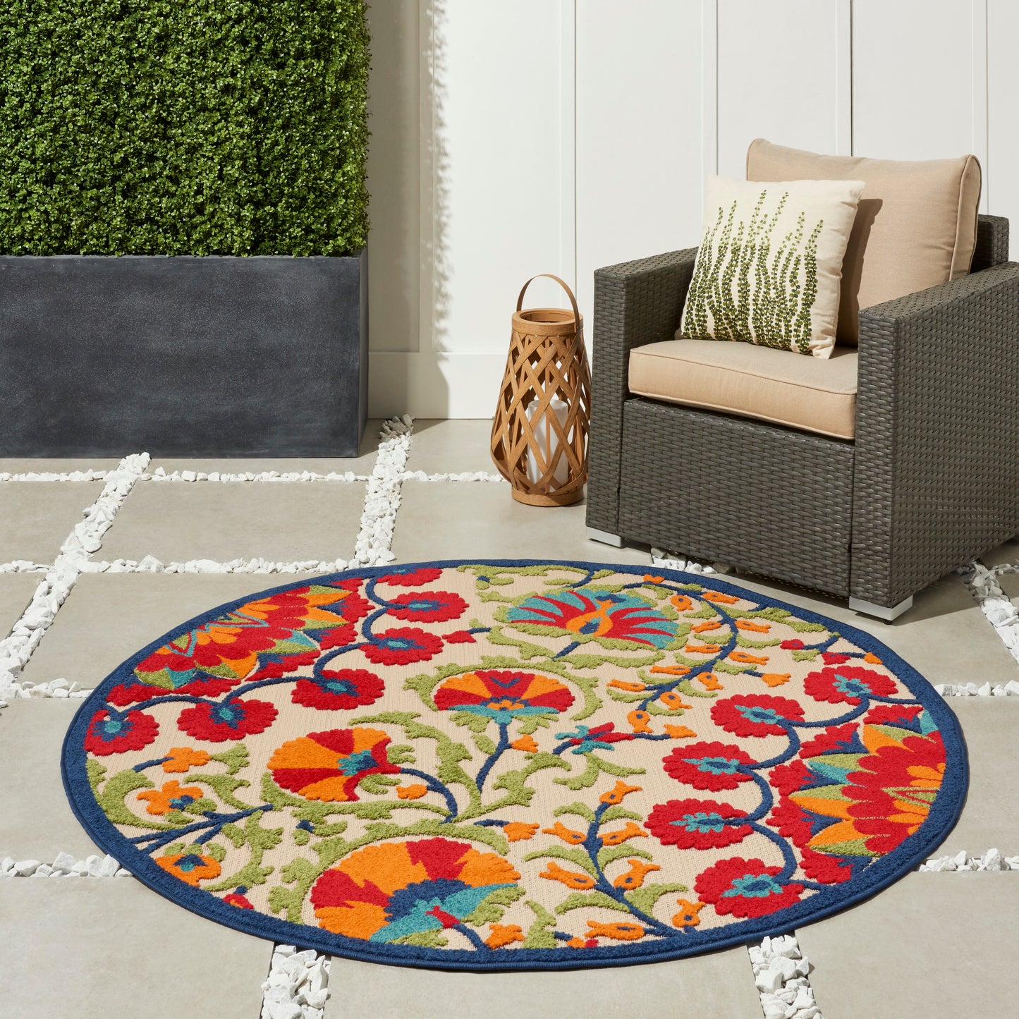 Round outdoor rug with a vibrant multicolor floral pattern and blue border on a stone patio beside a wicker armchair.